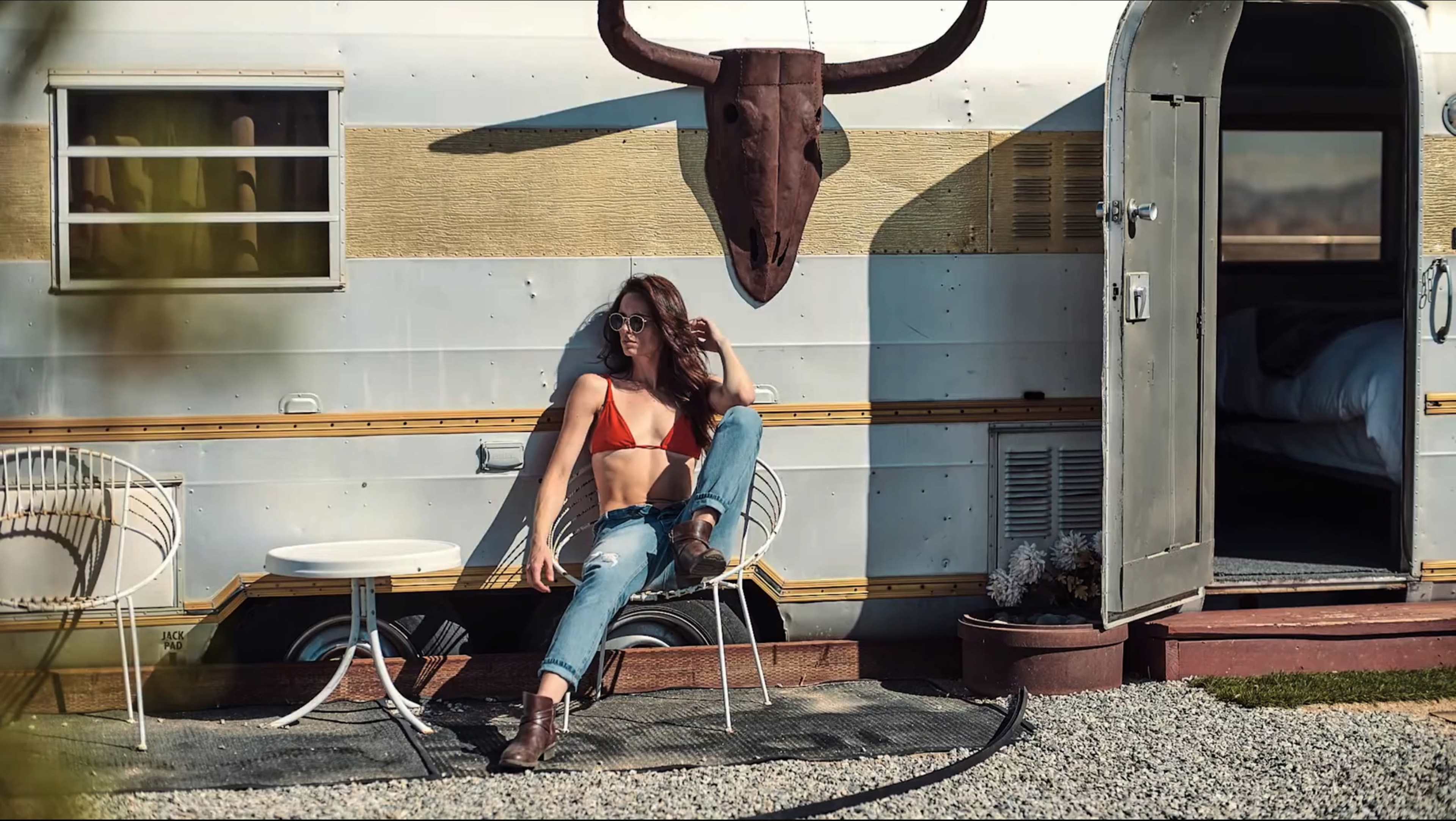 A woman in a red bikini top and jeans relaxes in a chair beside a vintage trailer adorned with a bull skull.