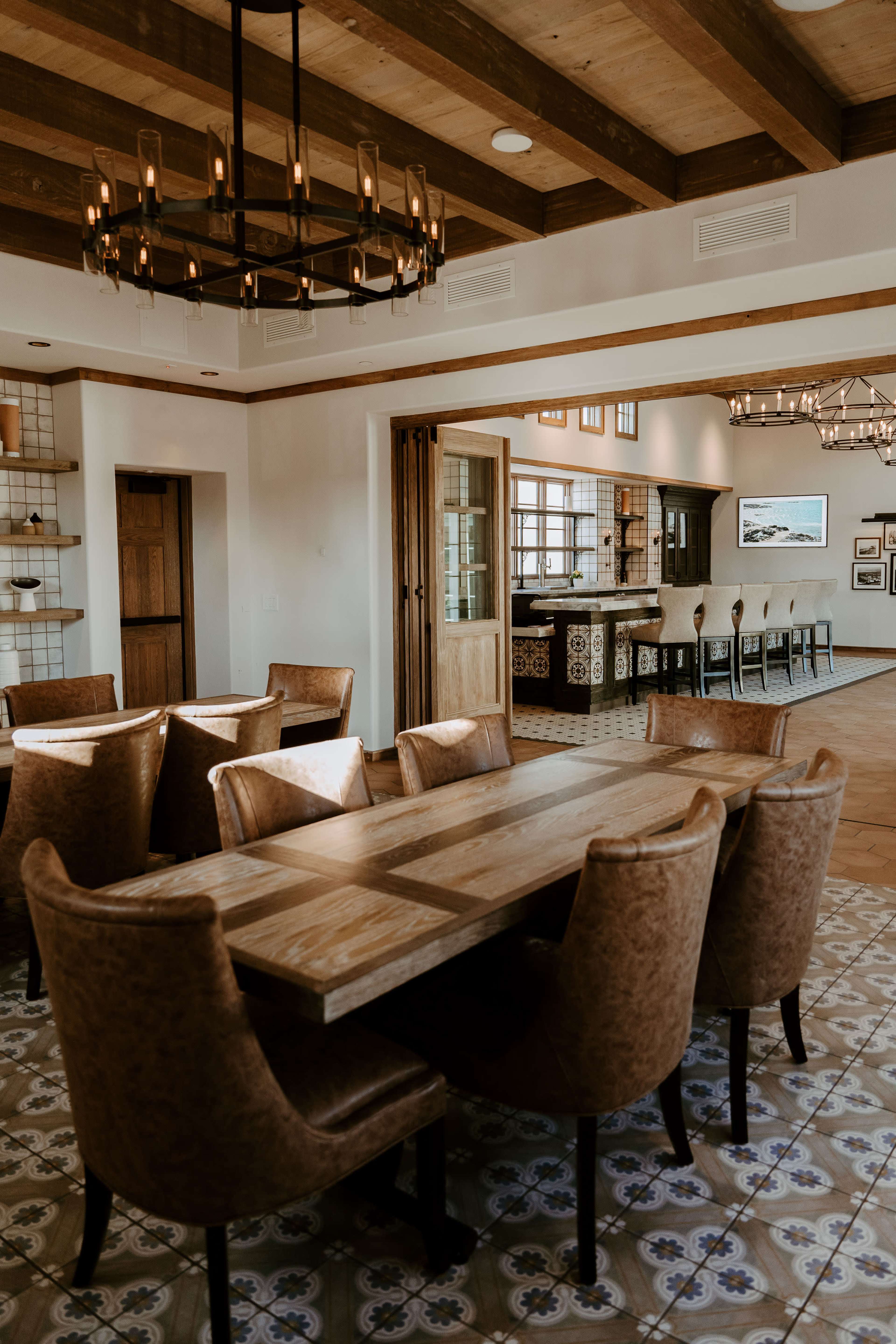 A dining area featuring a long wooden table surrounded by upholstered chairs, with a view of an adjacent kitchen area in a well-lit, modern interior.