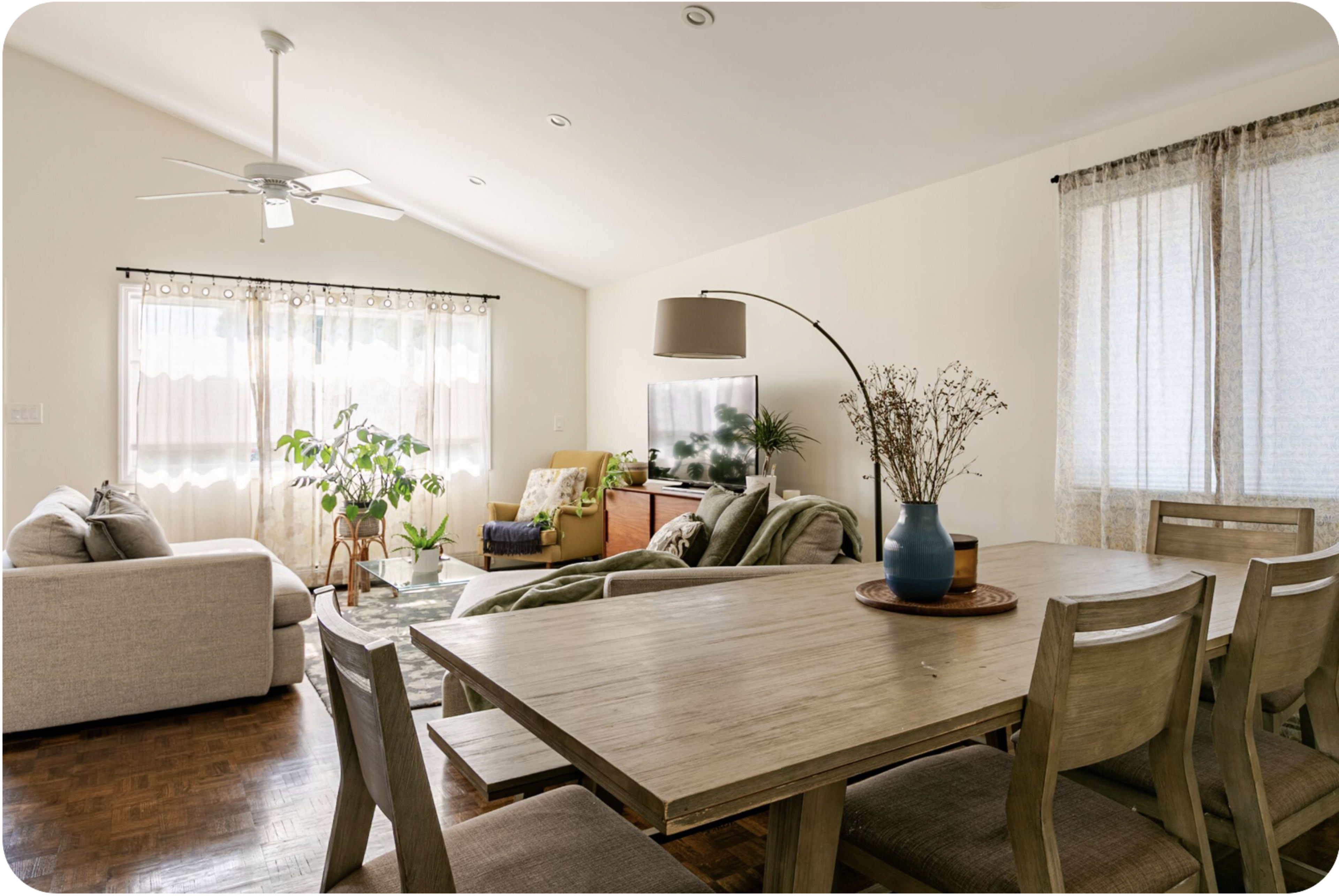 The image shows a bright living space featuring a dining table, a sofa, and a television surrounded by plants and natural light.
