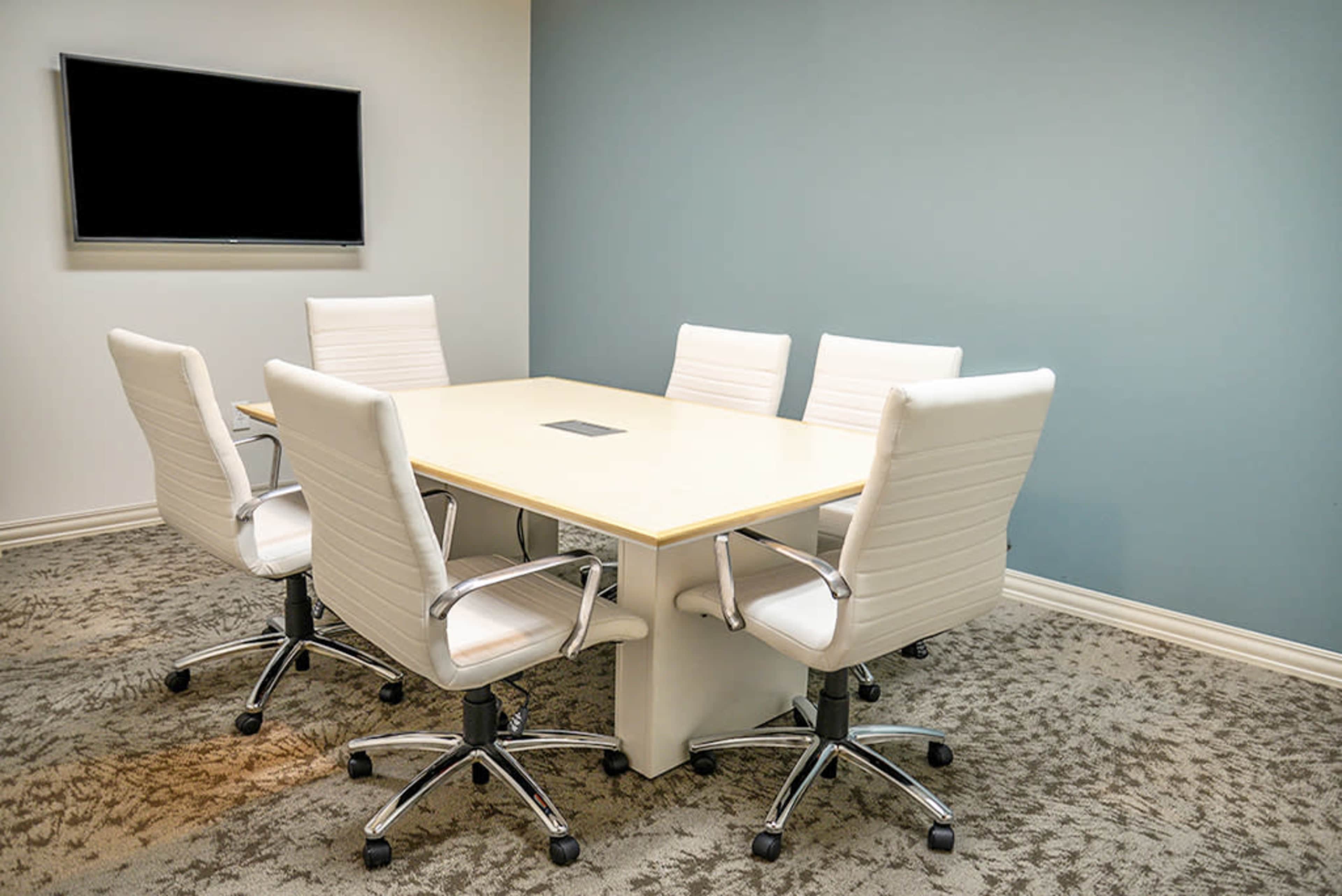 A conference room features a rectangular table surrounded by five white chairs and a wall-mounted screen.