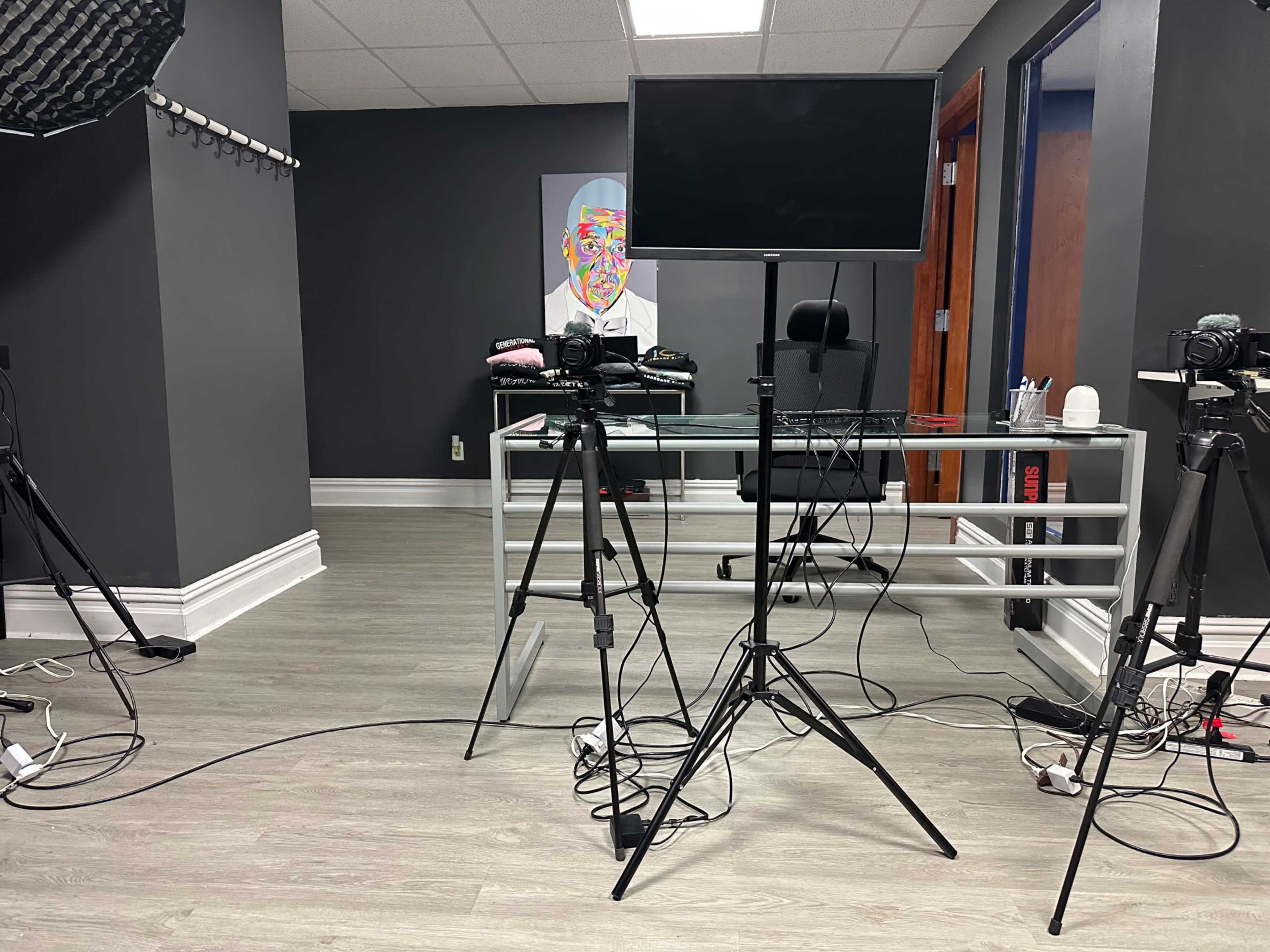A modern office setup with a glass desk, a monitor on a stand, and several cameras positioned on tripods around the room.