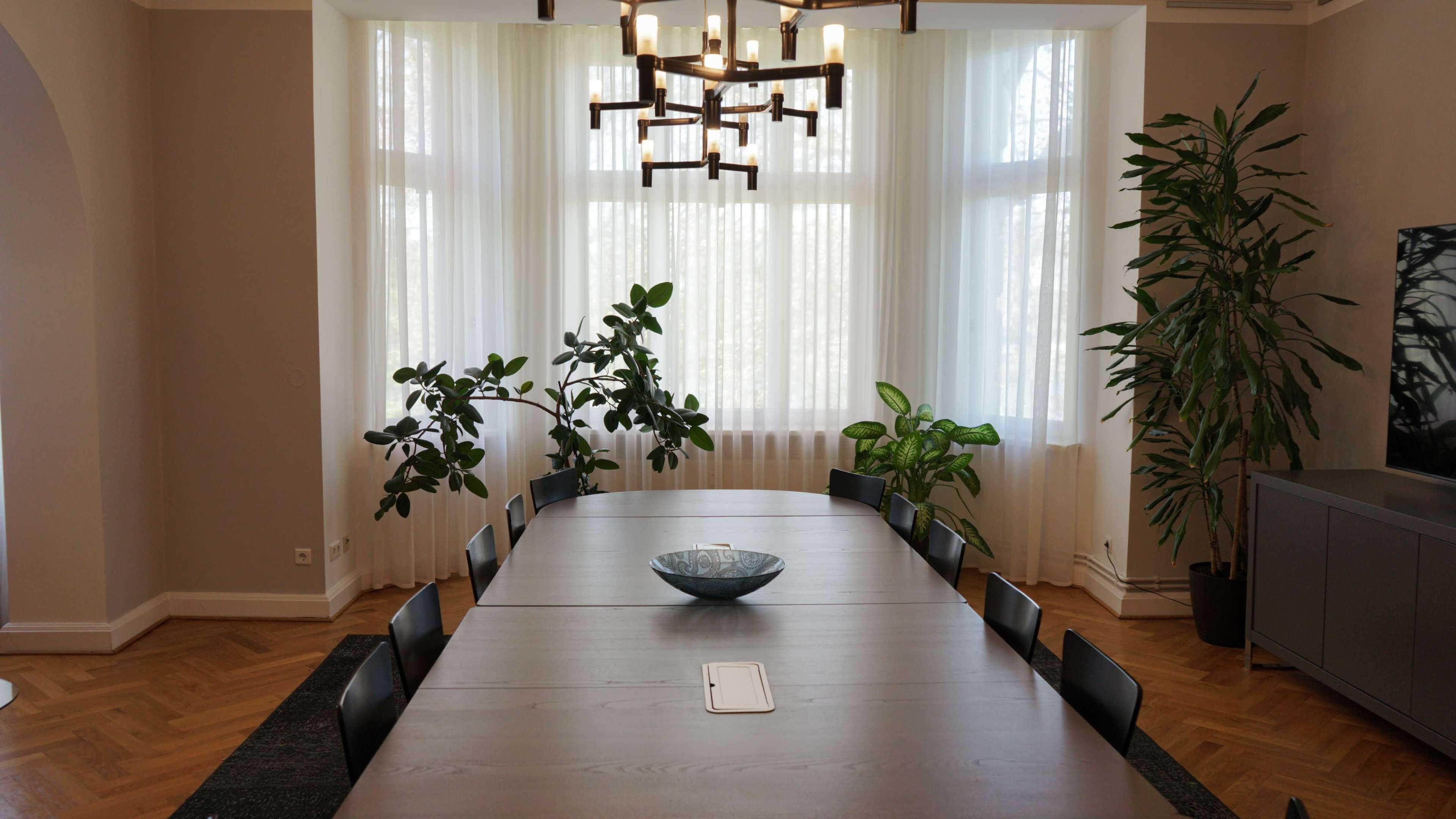 A long wooden dining table is set in a bright room with large windows, flanked by potted plants and a modern chandelier above.