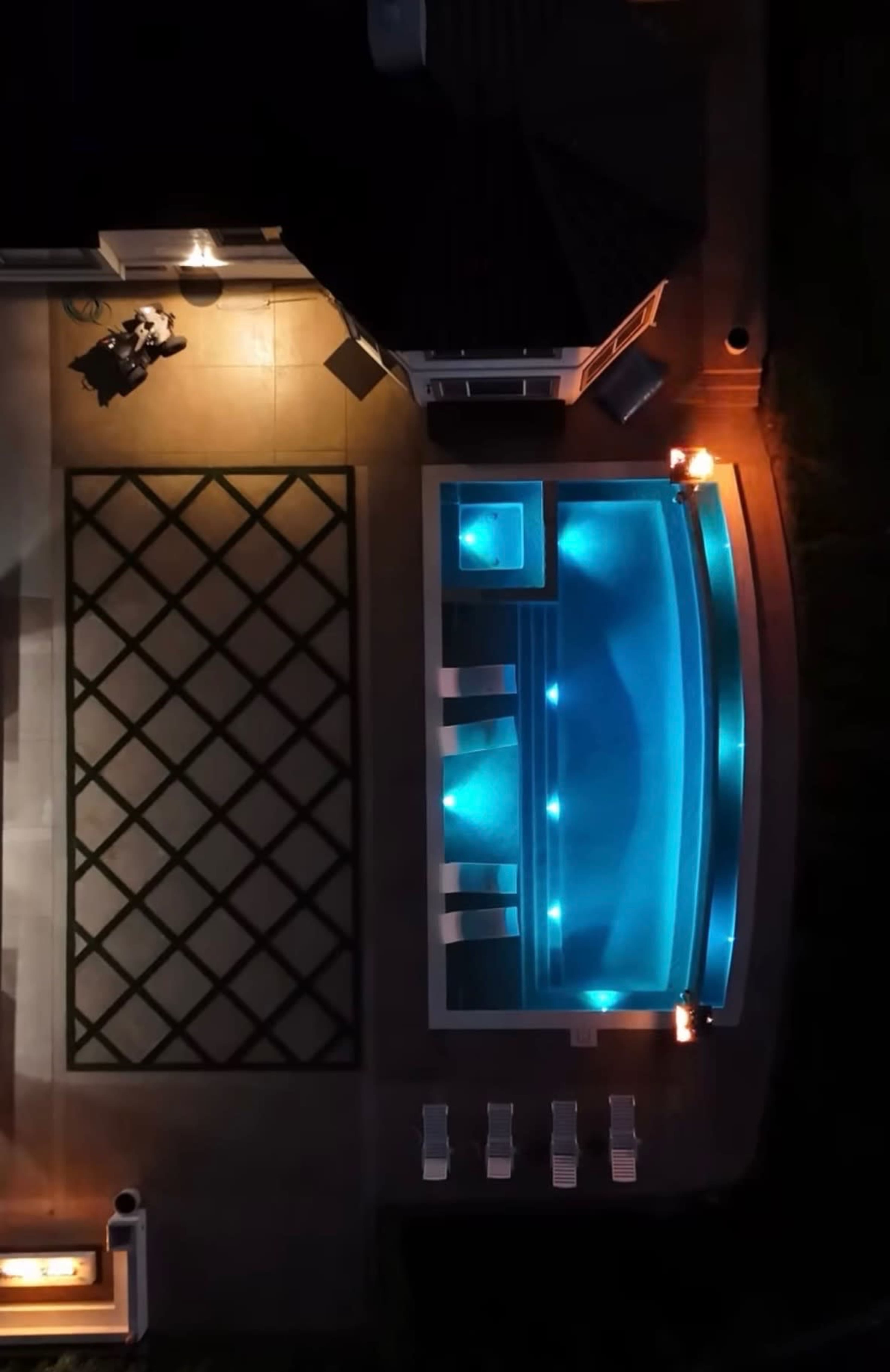 An aerial view of a swimming pool with illuminated water and surrounding lounge chairs, adjacent to a patio area with a geometric pattern.