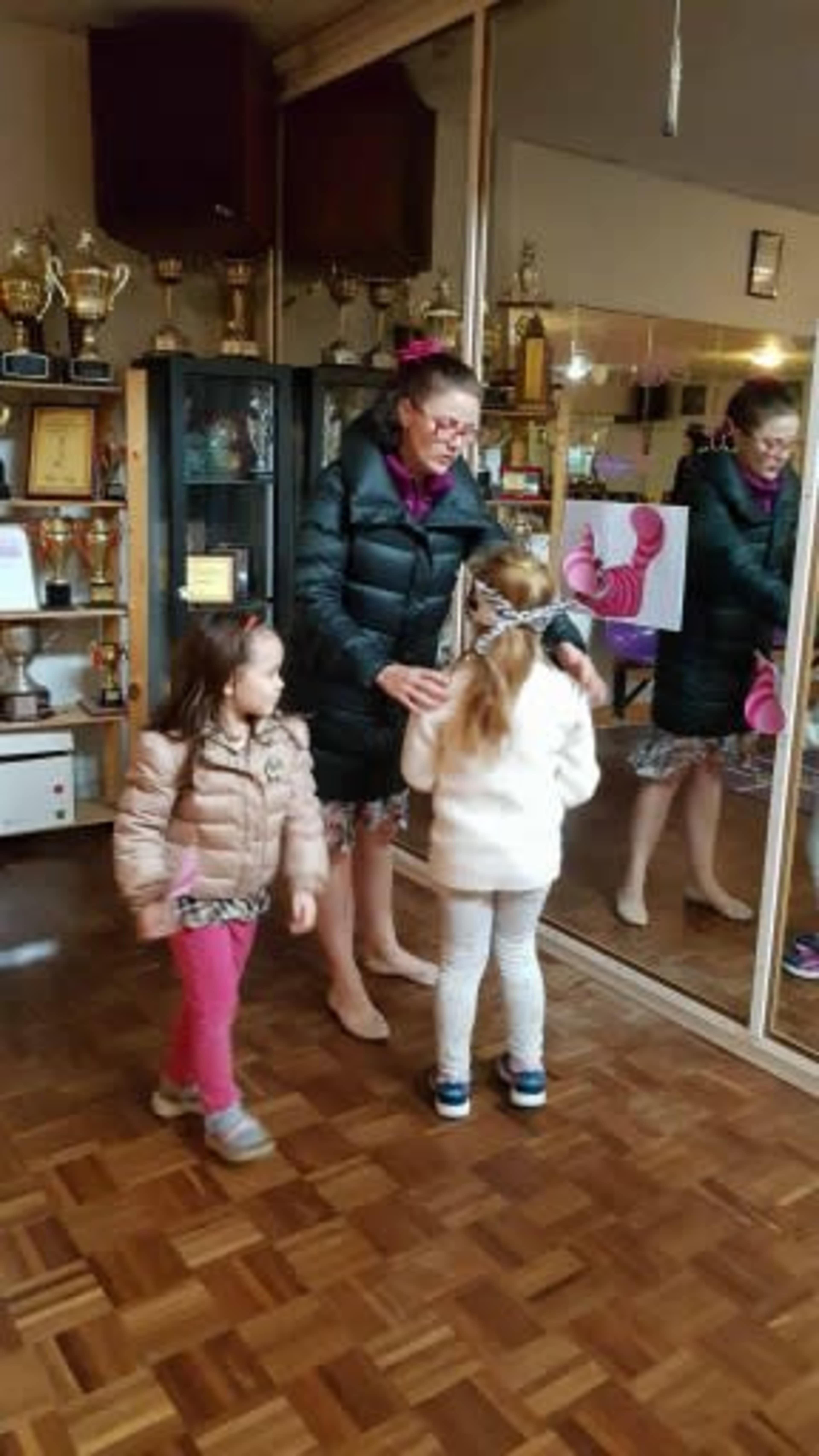 Two girls interacting with a woman in a room filled with trophies and awards, with a mirrored wall reflecting the scene.