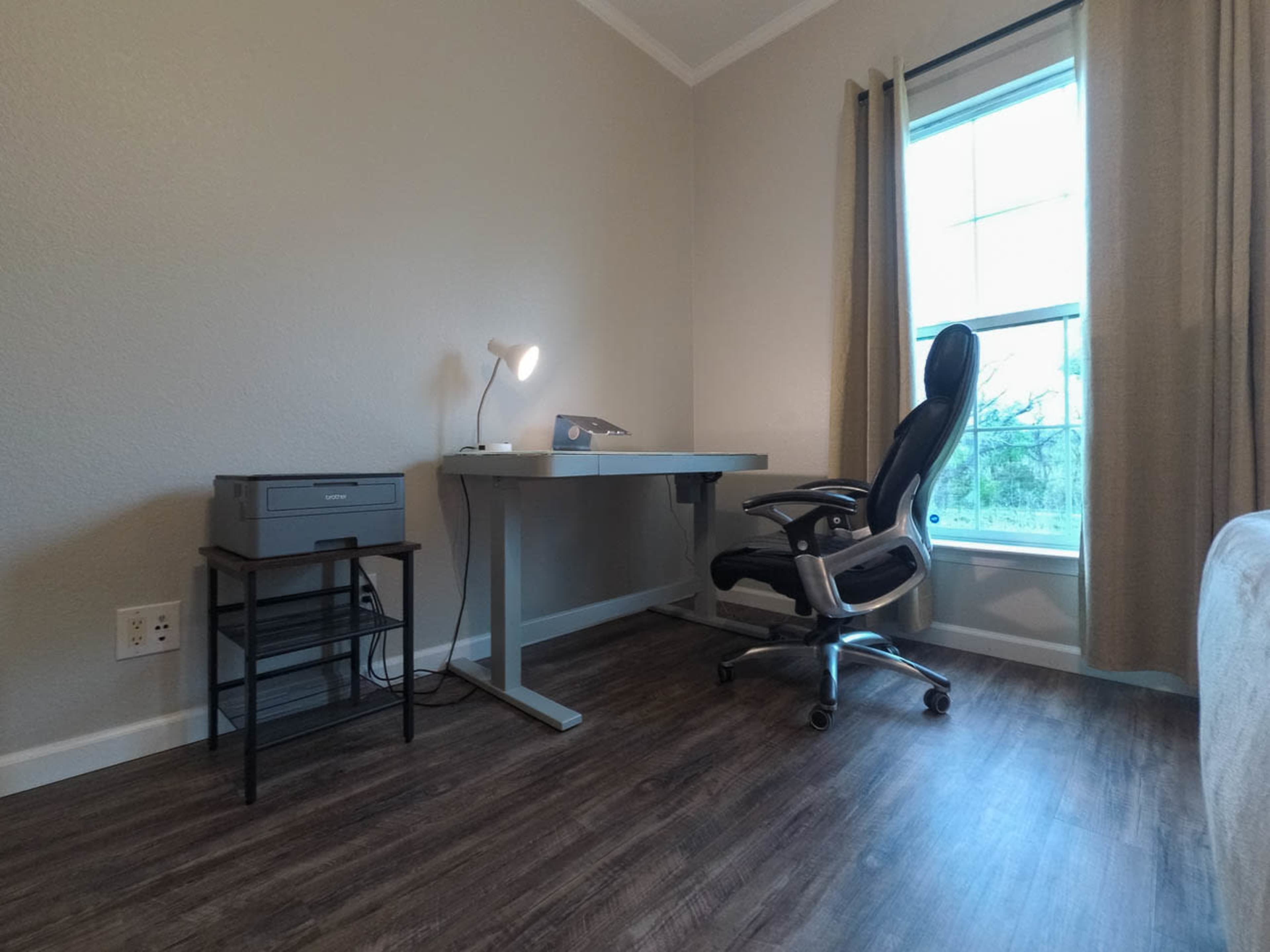 A minimalist home office setup featuring a desk, an office chair, a printer, and a window with curtains.