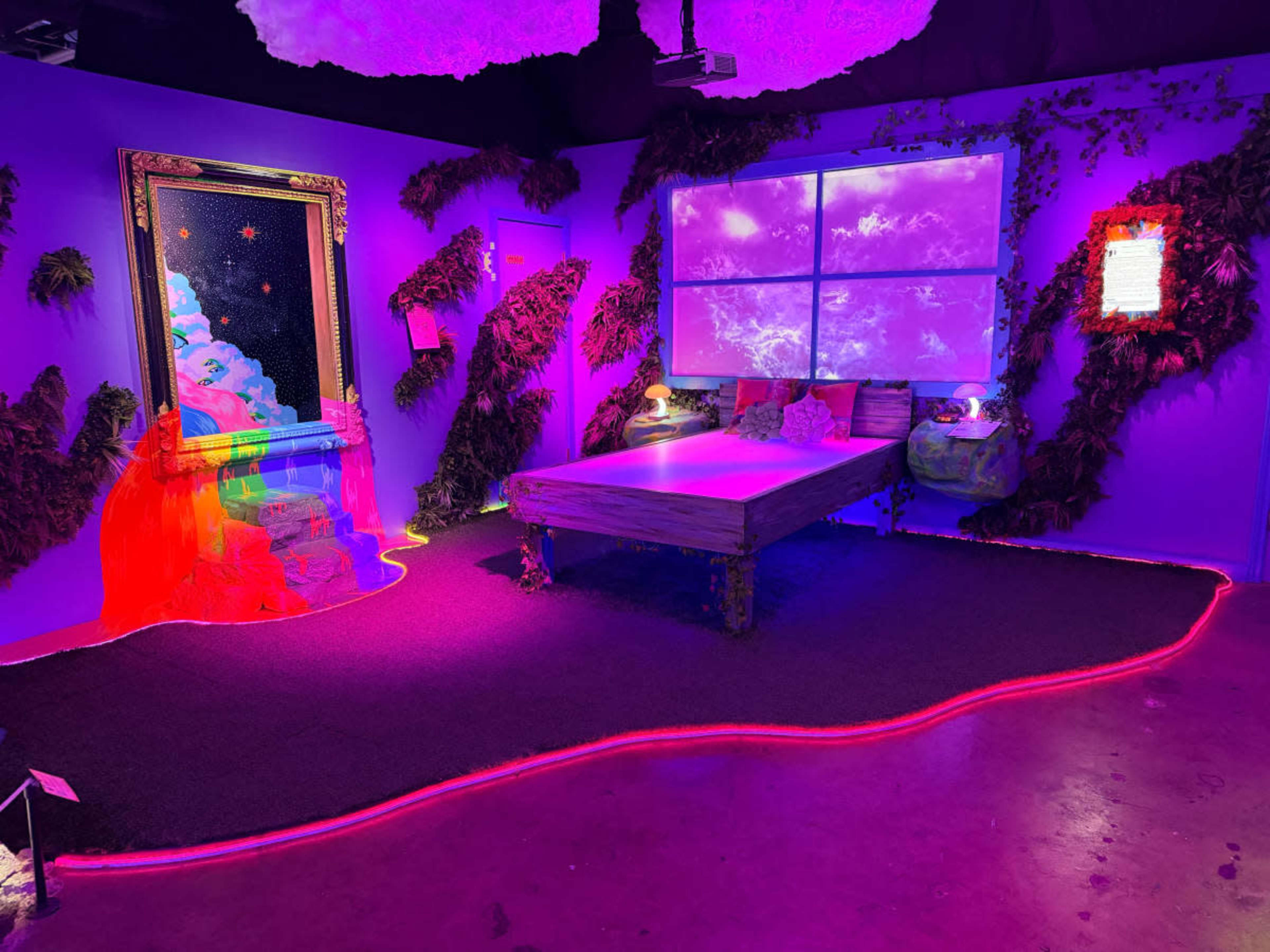 The image depicts a vibrant art installation featuring a bed surrounded by colorful lighting and decorative plants, with a large framed artwork on the wall.