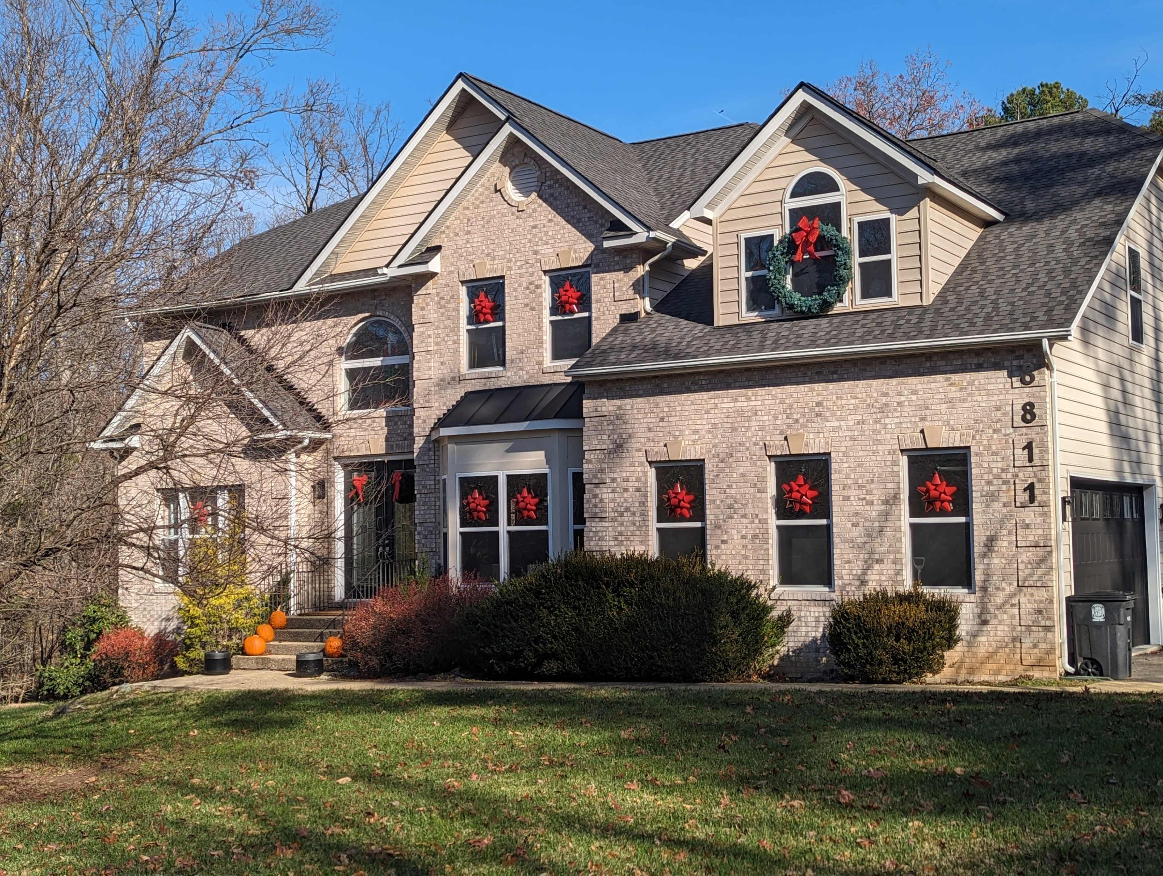 A large brick house displays festive decorations, including red snowflake ornaments in the windows and a wreath on the front door, set against a clear blue sky.