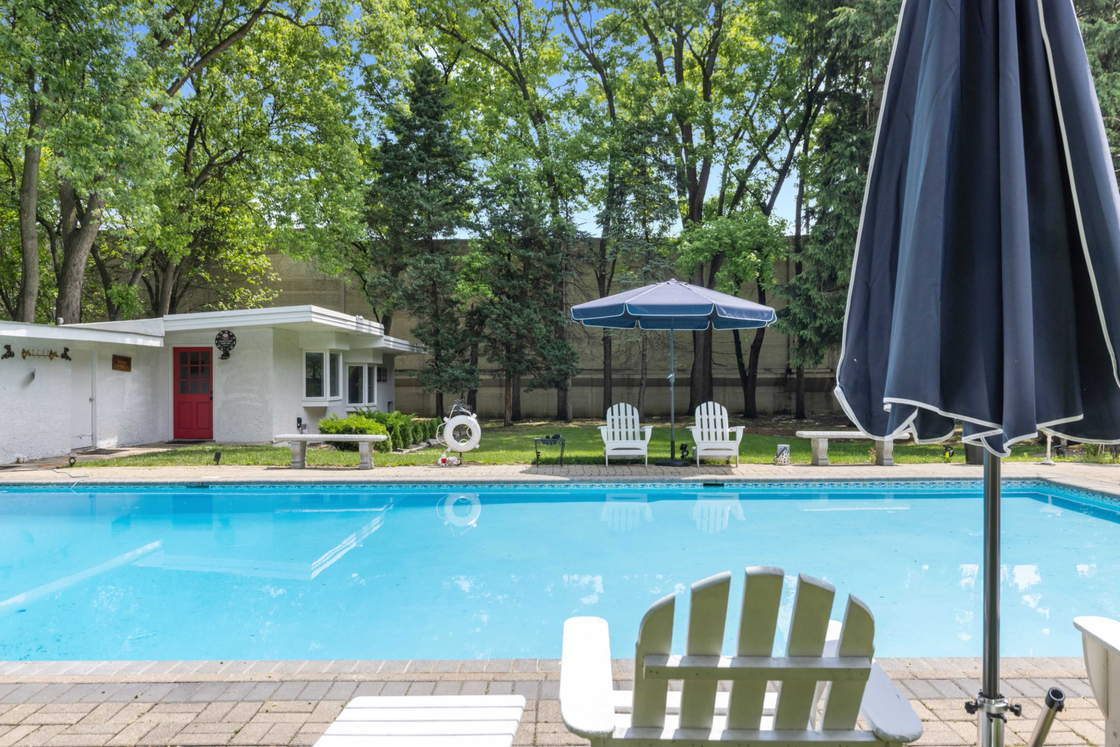 A clear swimming pool is surrounded by green trees, lounge chairs, and an umbrella, with a white exterior building visible in the background.
