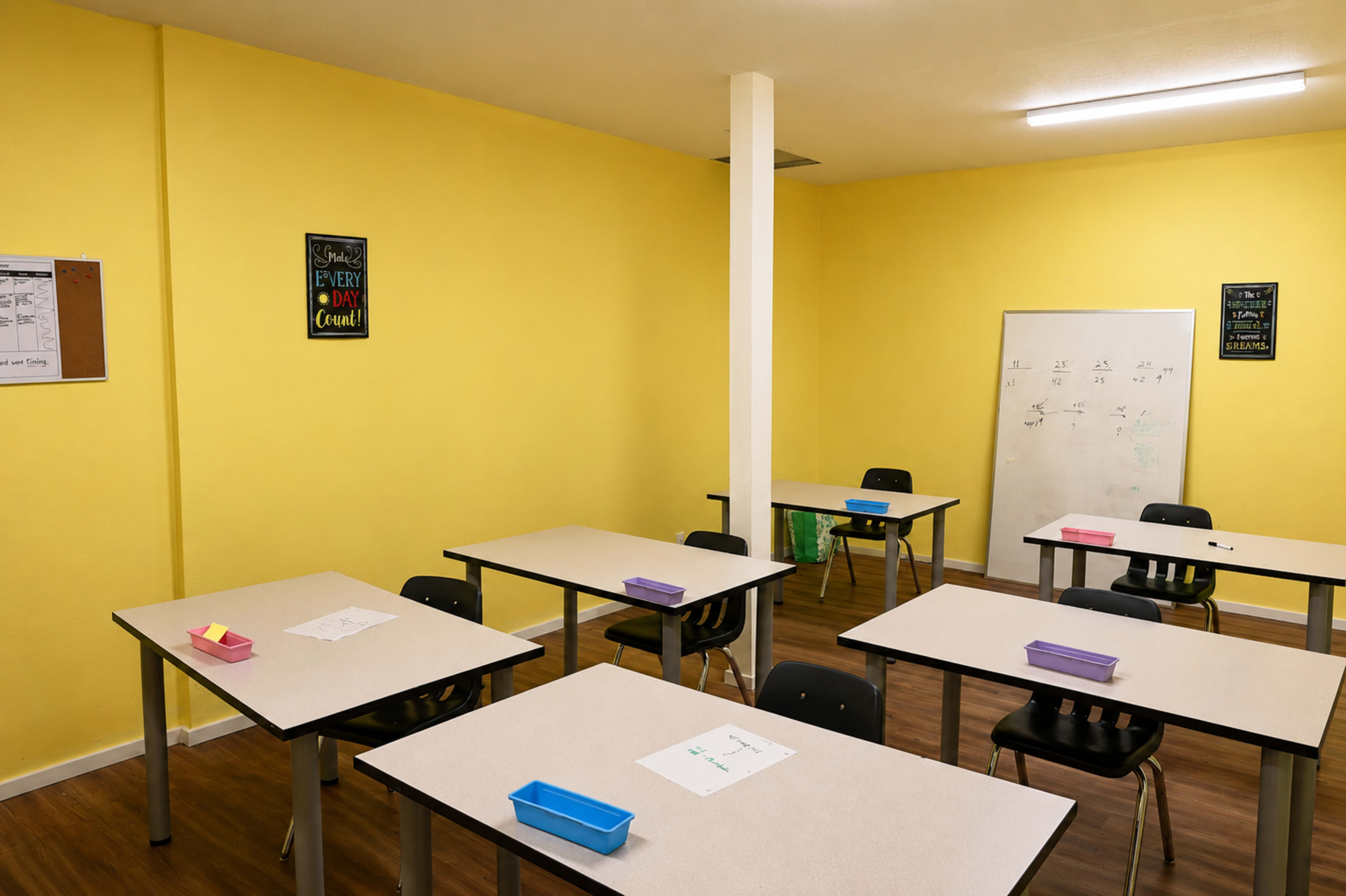 The image shows a classroom with yellow walls, several tables arranged with chairs, and a whiteboard on one side.