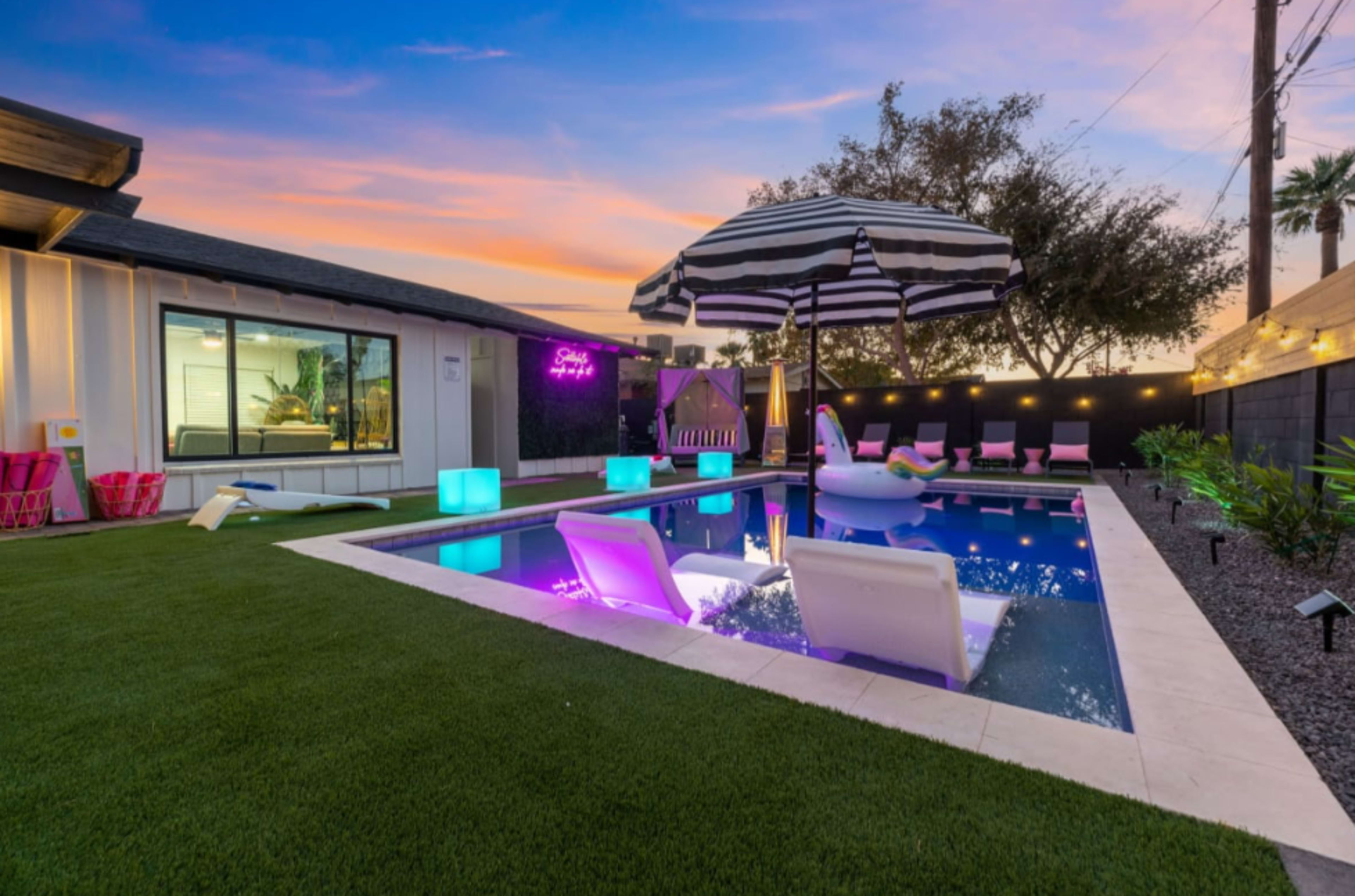 The image shows a modern backyard with a swimming pool, colorful illuminated furniture, and a large striped umbrella set against a sunset sky.
