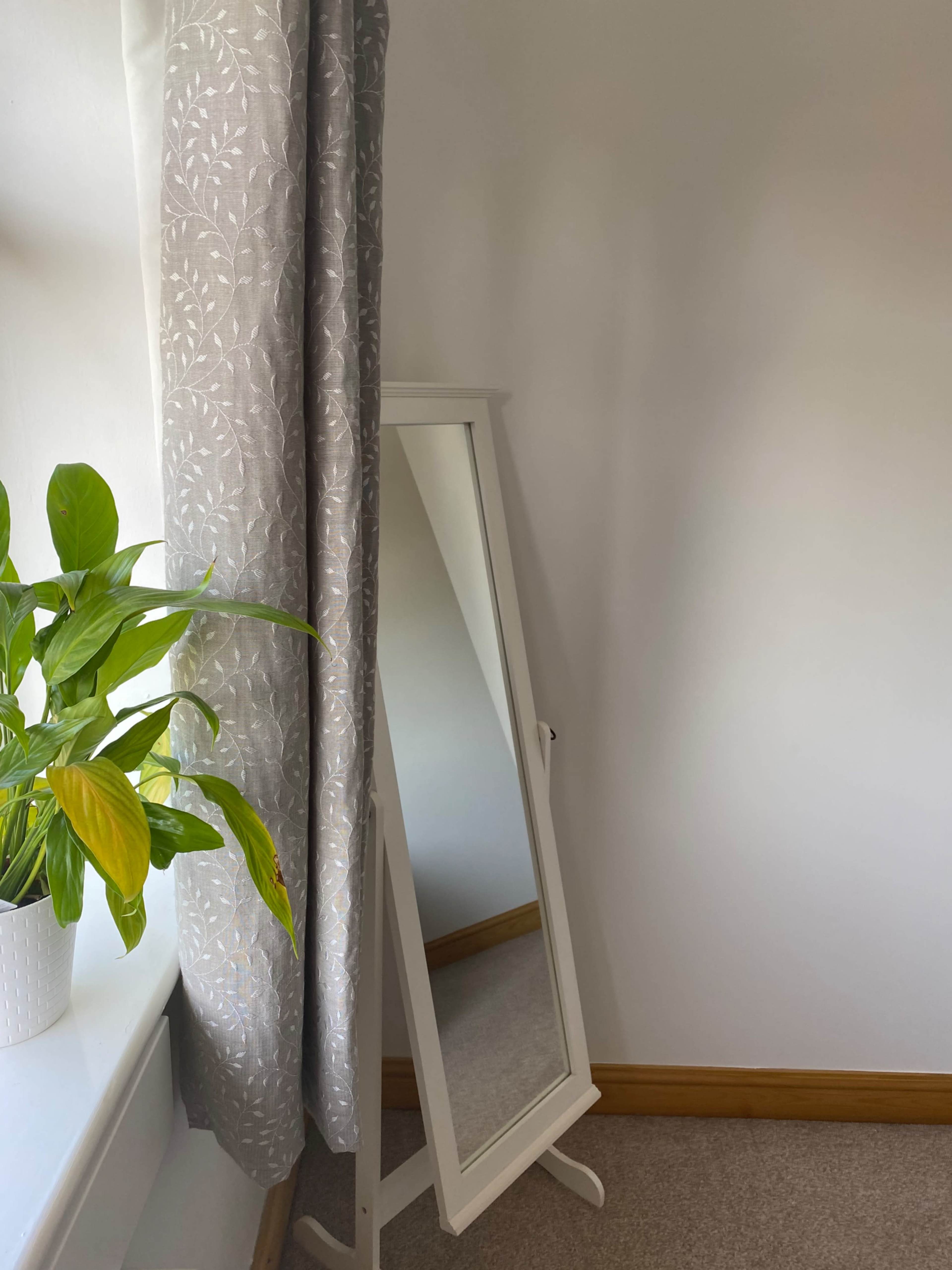 A standing mirror is positioned beside a window with a potted plant on the windowsill and gray curtain draping nearby.