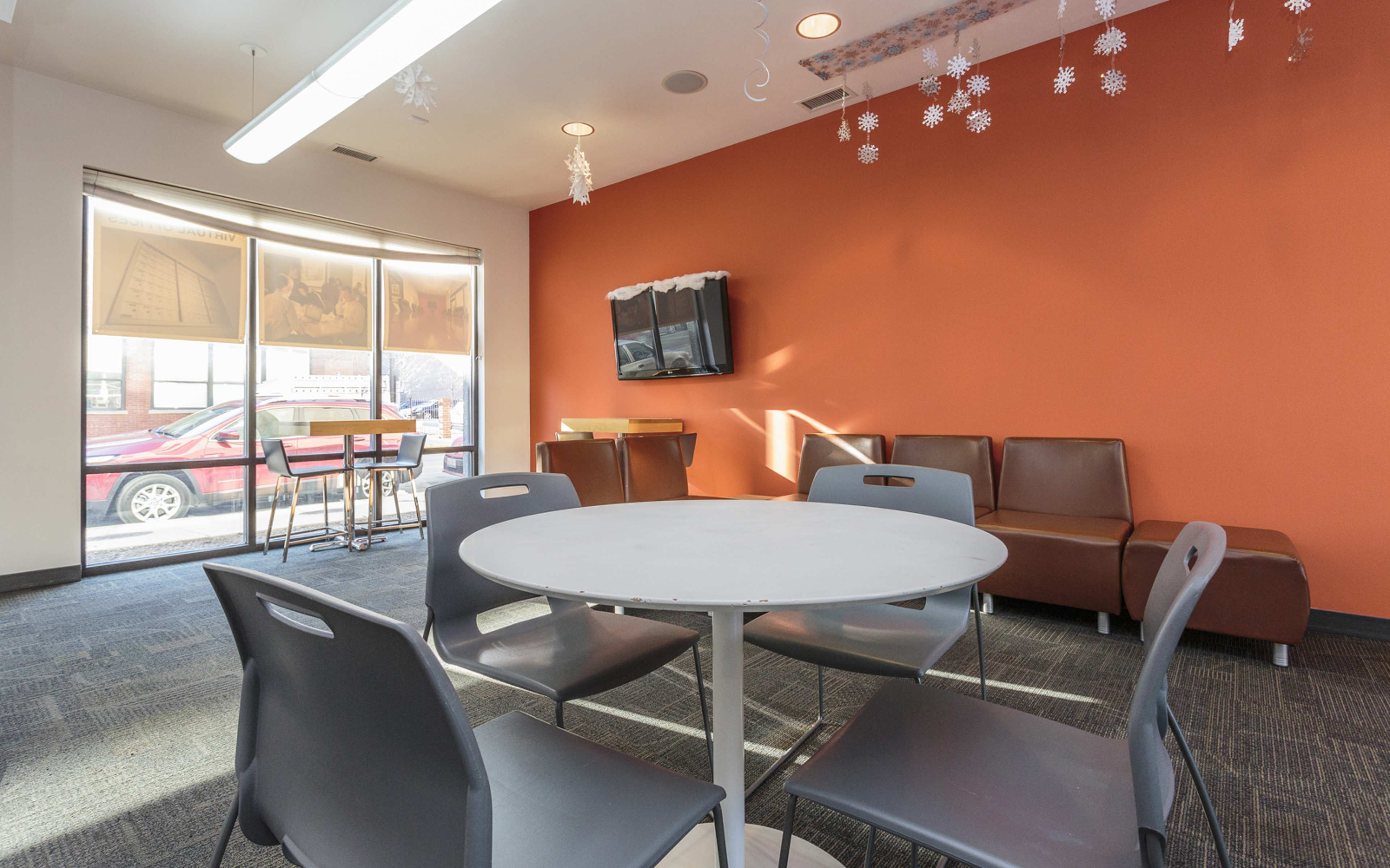 The image shows a modern communal space featuring a round table with several chairs, a wall-mounted television, and brown seating along an orange wall.