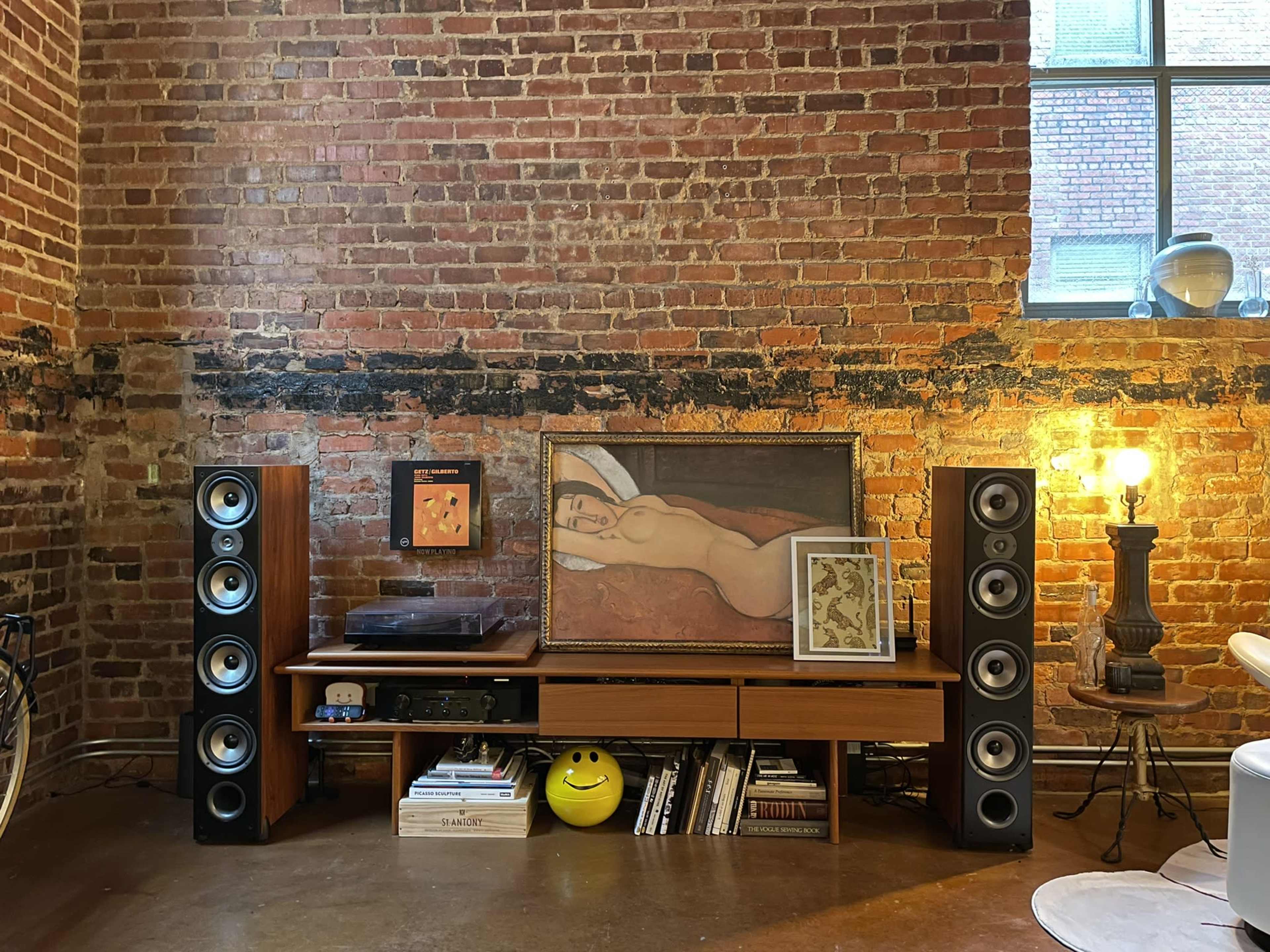 A wooden media console with tall speakers on either side displays a large painting and a framed artwork, against a backdrop of exposed brick walls and a window.