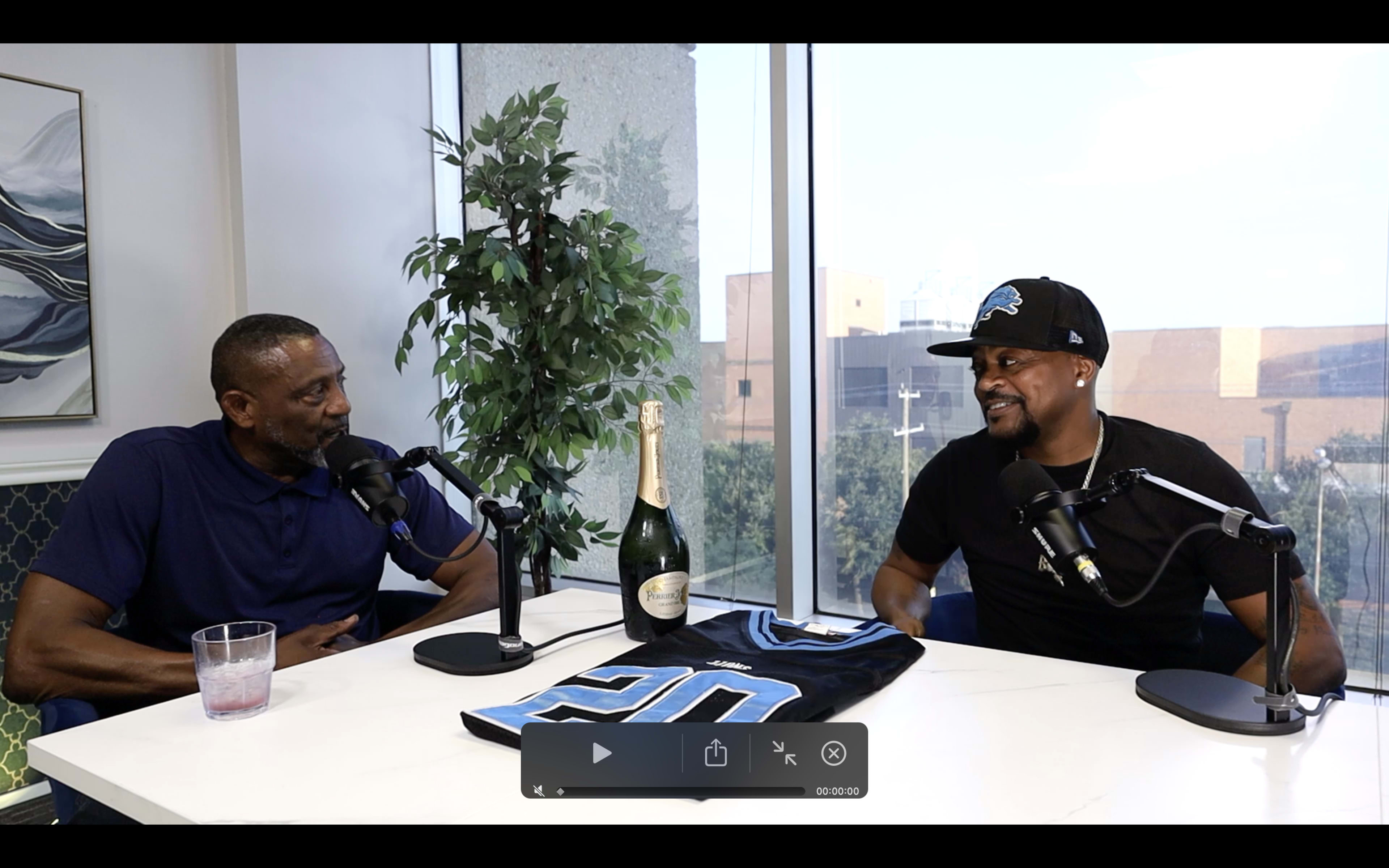 Two men sit at a white table with microphones, engaging in conversation, while a bottle of champagne and a blue garment are positioned in front of them.