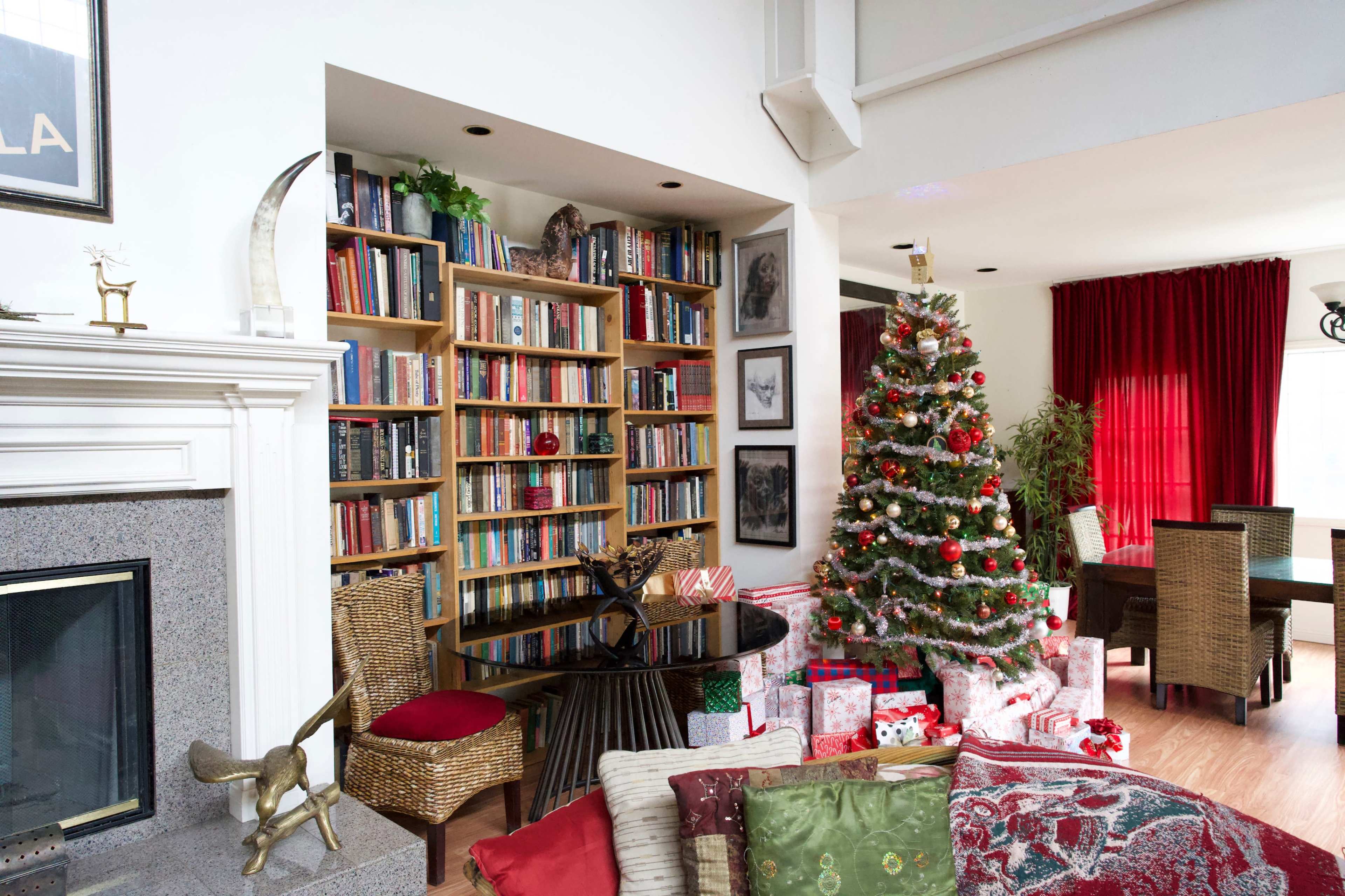 A cozy living room features a large bookshelf filled with books, a decorated Christmas tree surrounded by wrapped presents, and a dining area in the background.