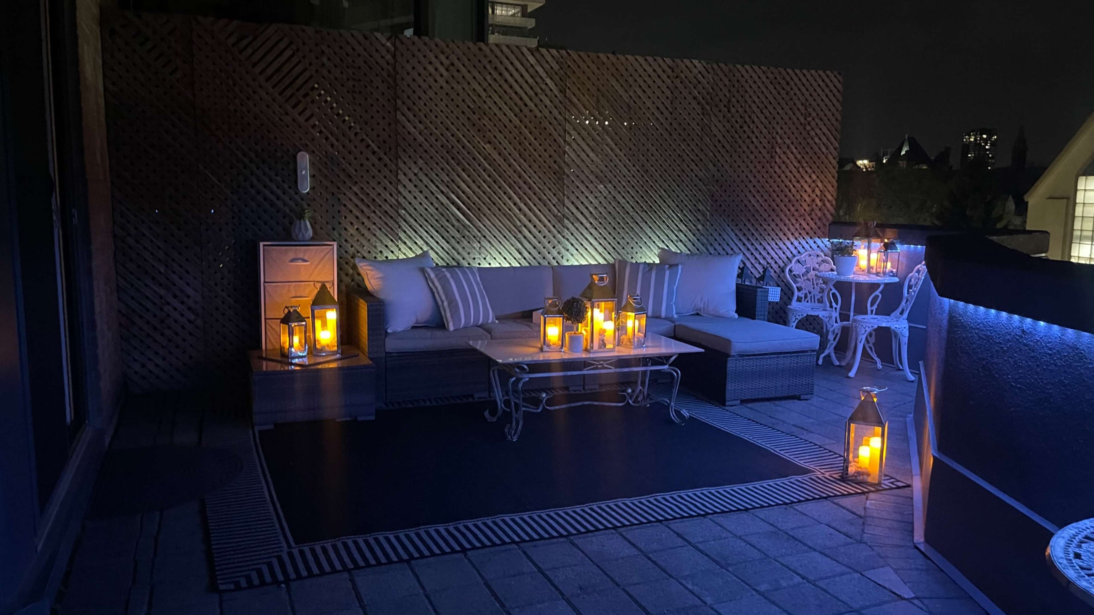A rooftop terrace features a seating area with white furniture, illuminated by several lanterns and soft blue lighting.