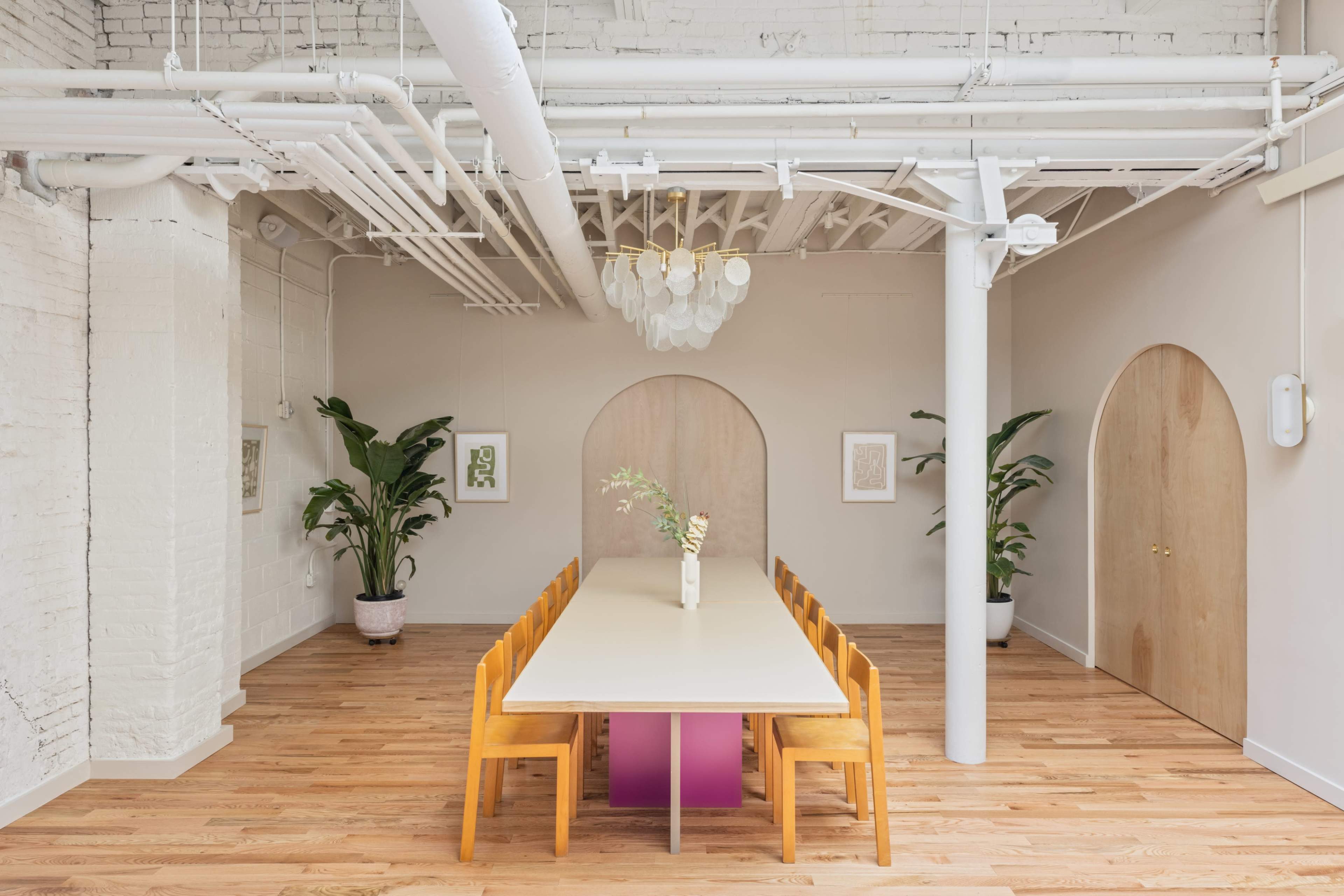 A long table surrounded by wooden chairs is positioned in a bright, modern room with arched doorways and potted plants.