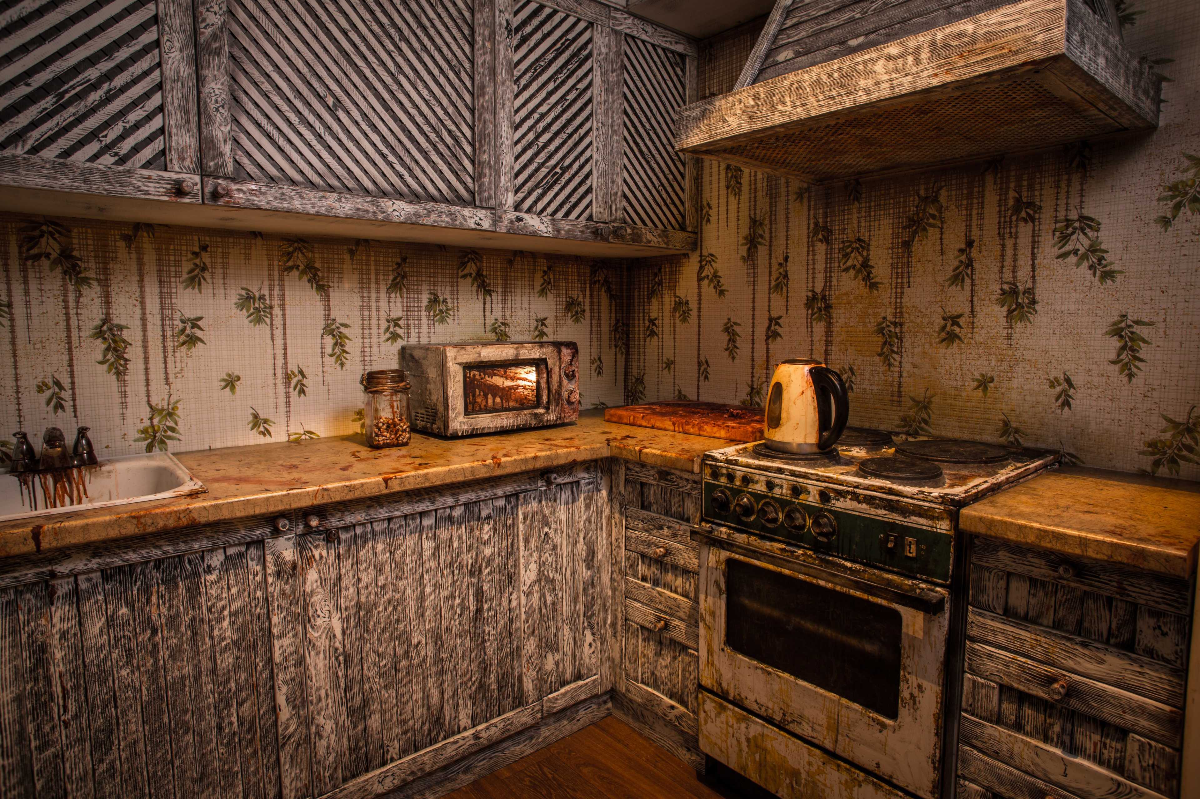 The image shows a weathered kitchen with a rustic wooden cabinet, an old stove, a microwave, and a kettle on a stained countertop.