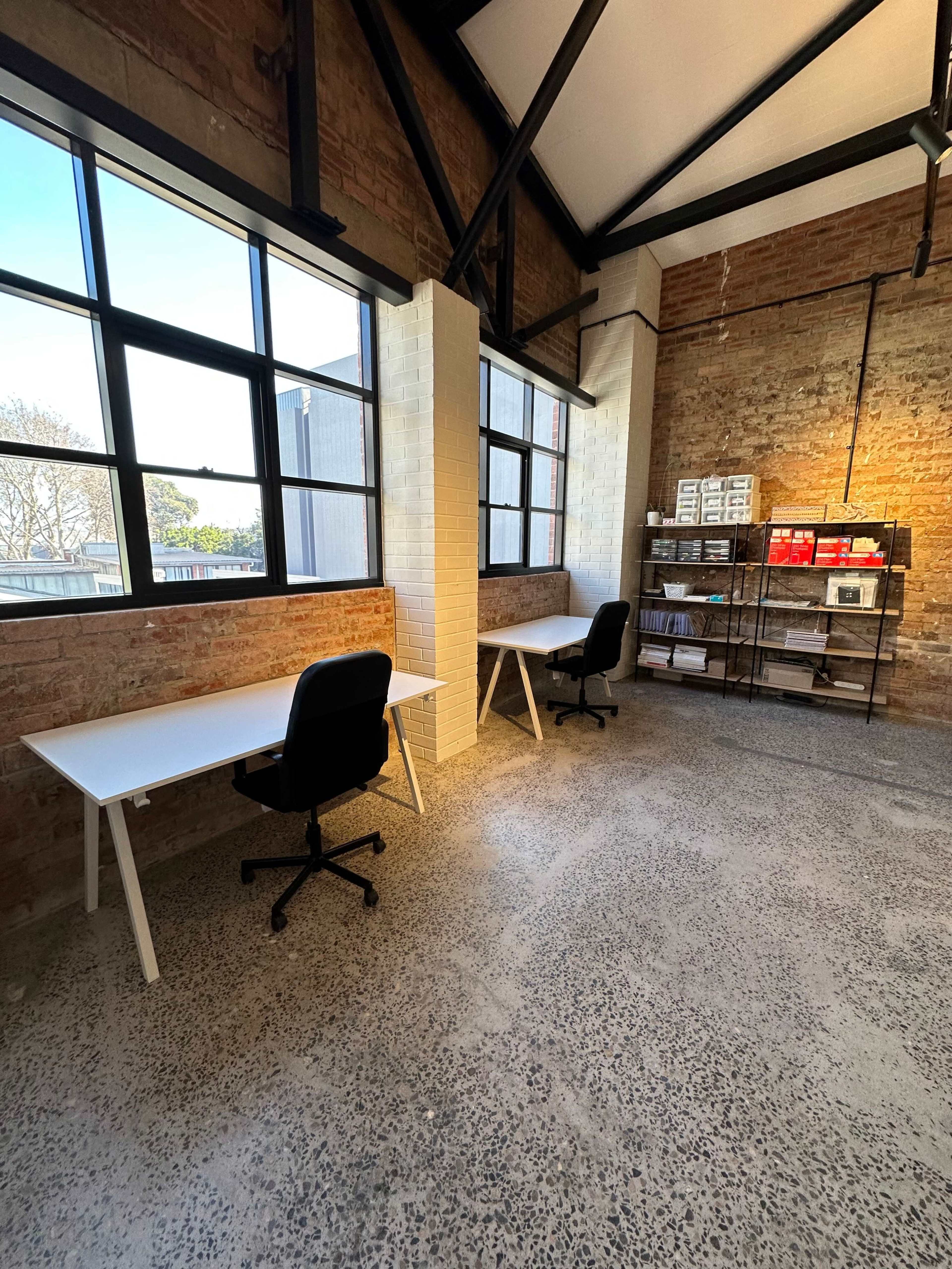 A spacious office area with two white desks and black chairs, illuminated by natural light from large windows, alongside a shelving unit containing various materials against a brick wall.
