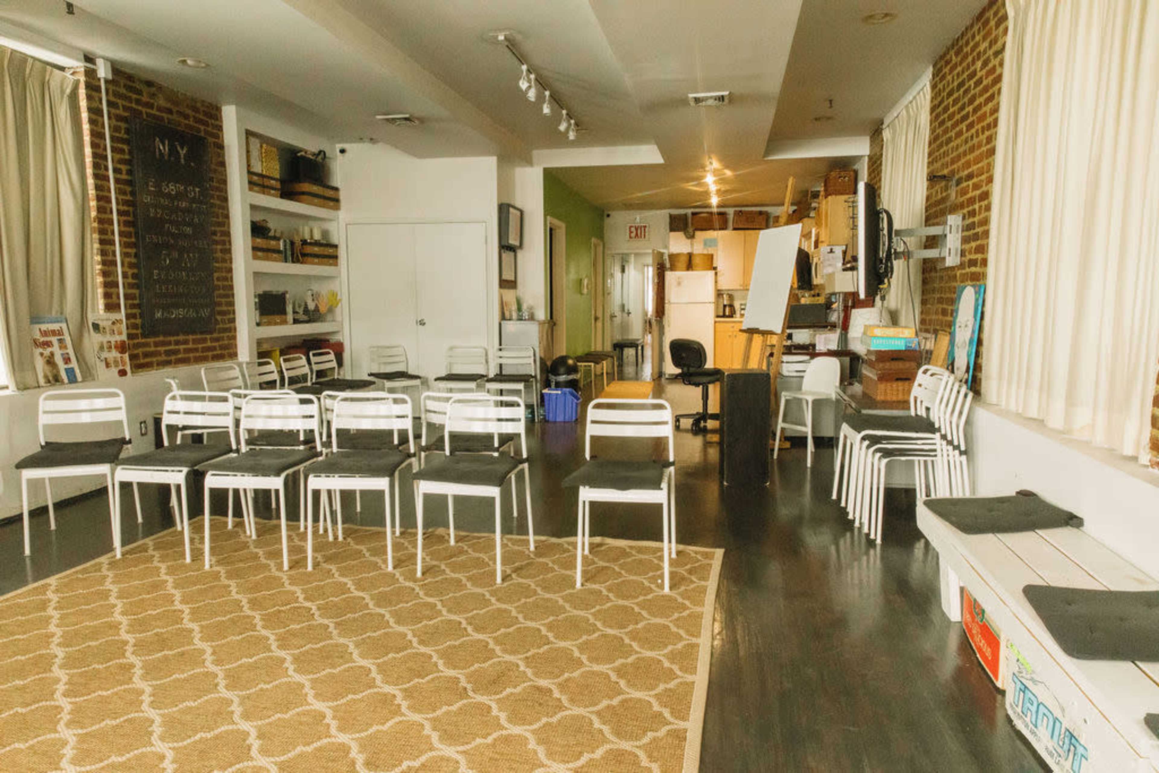 The image shows a spacious room with rows of white chairs facing a front area, wooden flooring, and large windows.