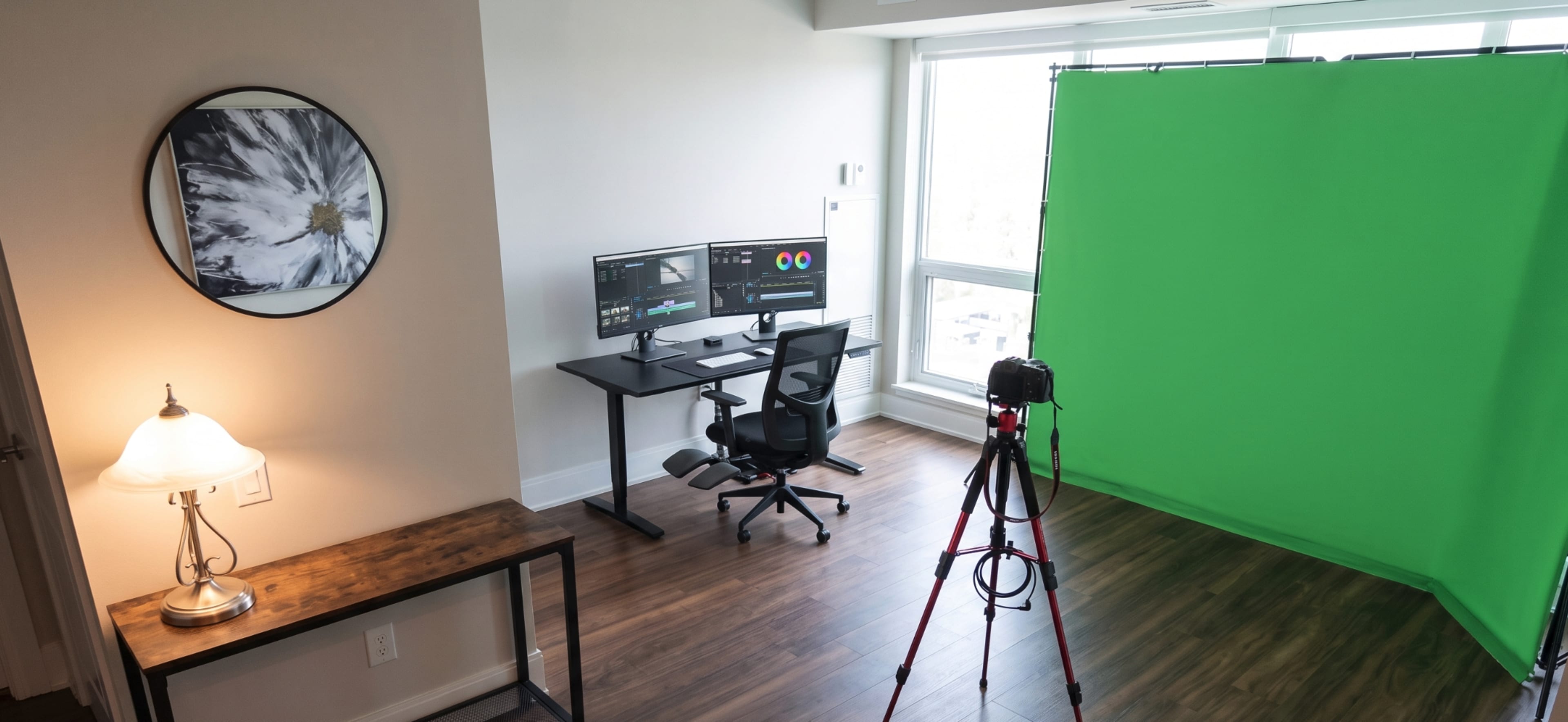 The image shows a room with a desk featuring dual monitors, a camera on a tripod, and a green backdrop for video production.