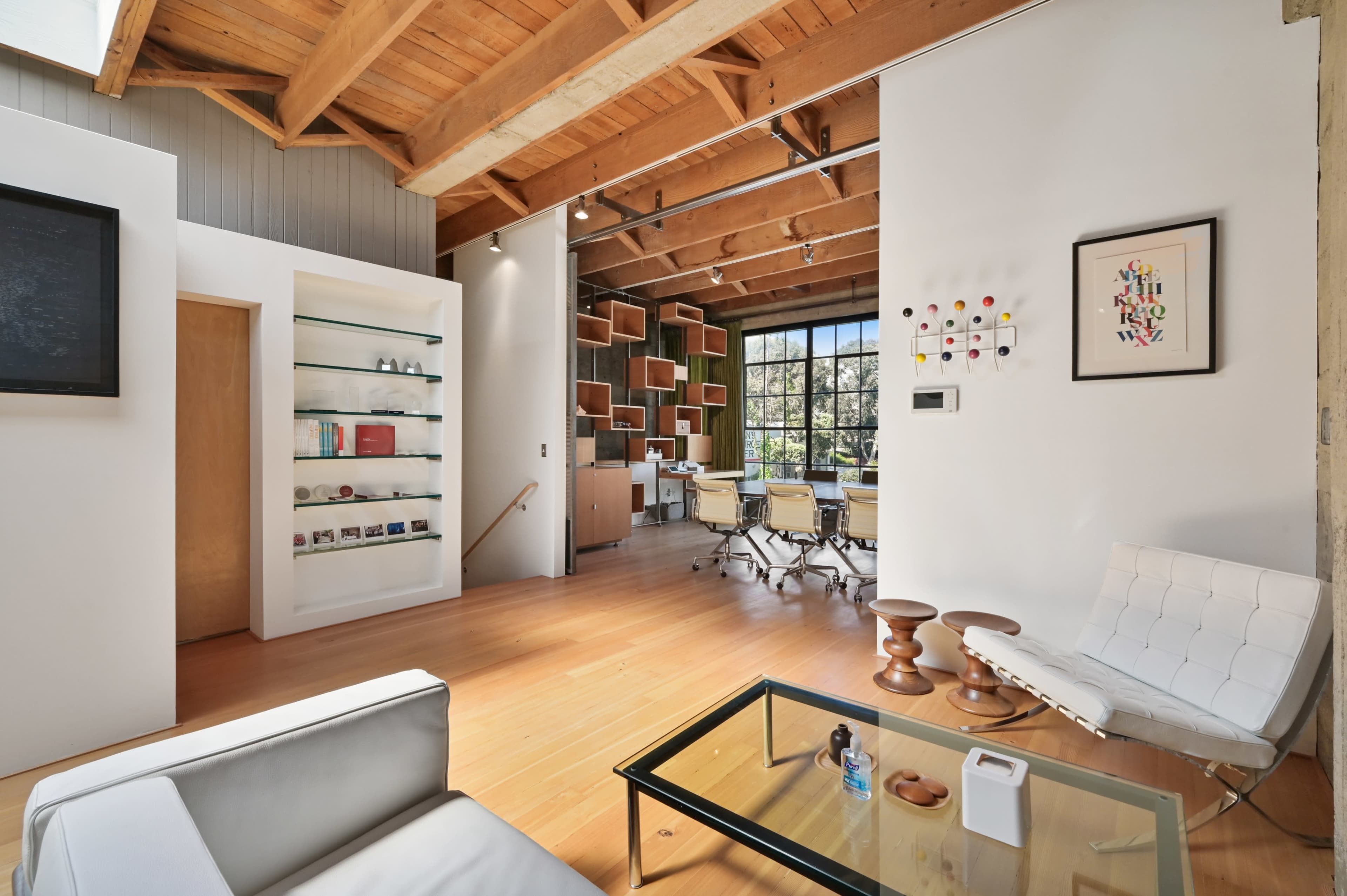 A modern office space with wooden beams, a glass coffee table, a white sofa, and a workstation surrounded by bookshelves.