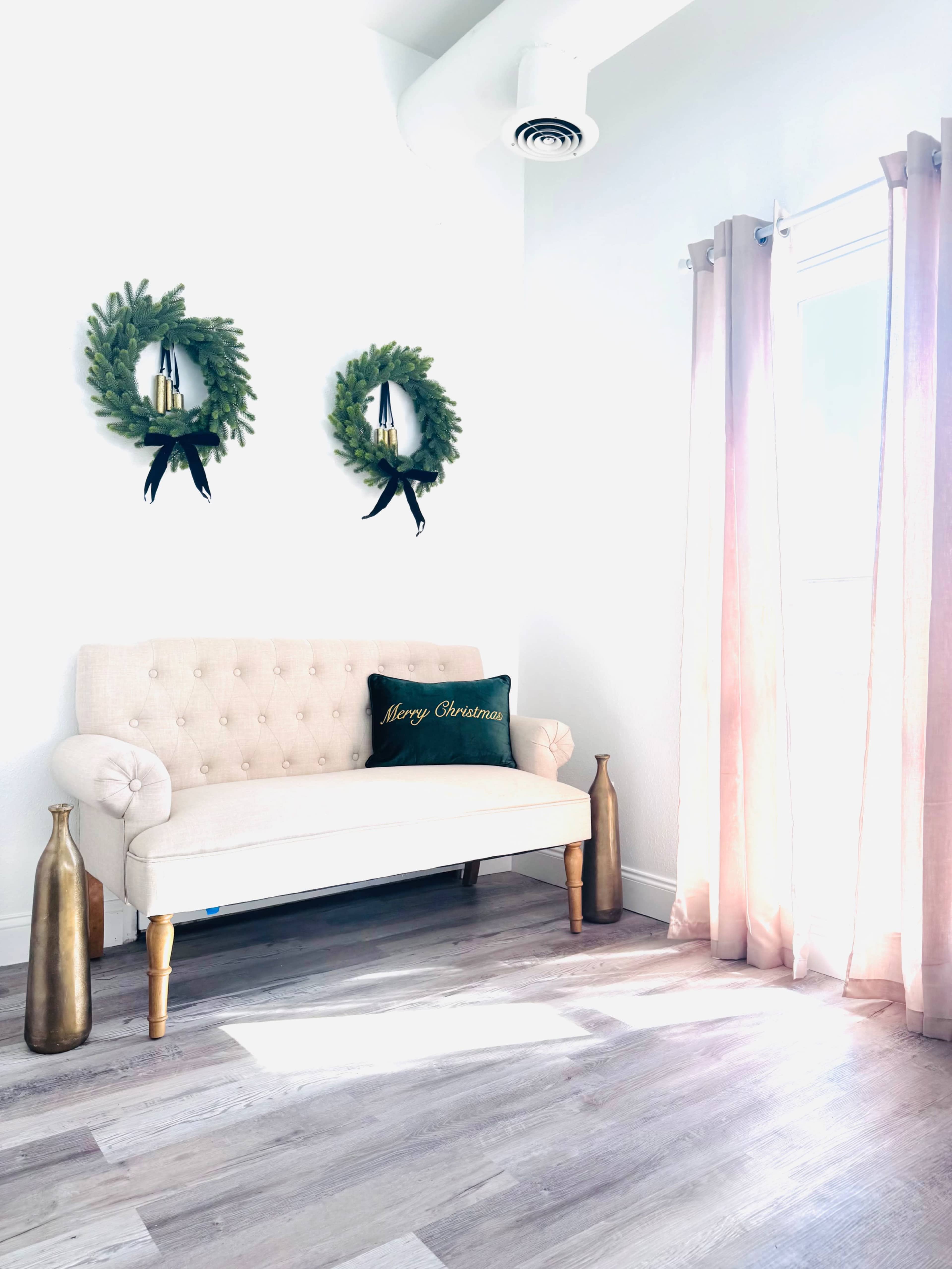 A light-filled room features a beige sofa with decorative pillows, a potted plant, and sheer pink curtains.