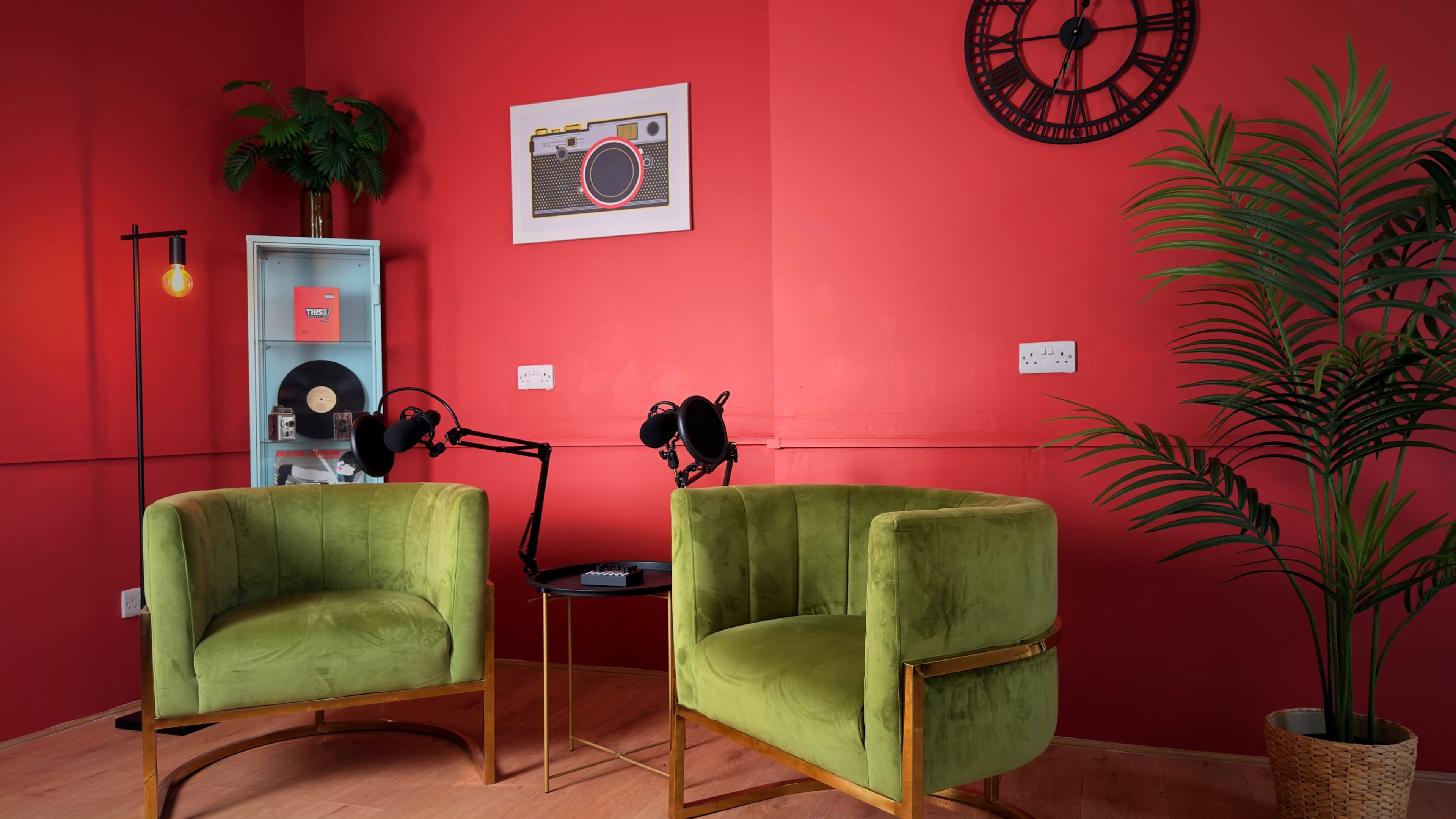 The image shows a vibrant room with two green chairs, a small round table, and a wall adorned with a clock and framed artwork.