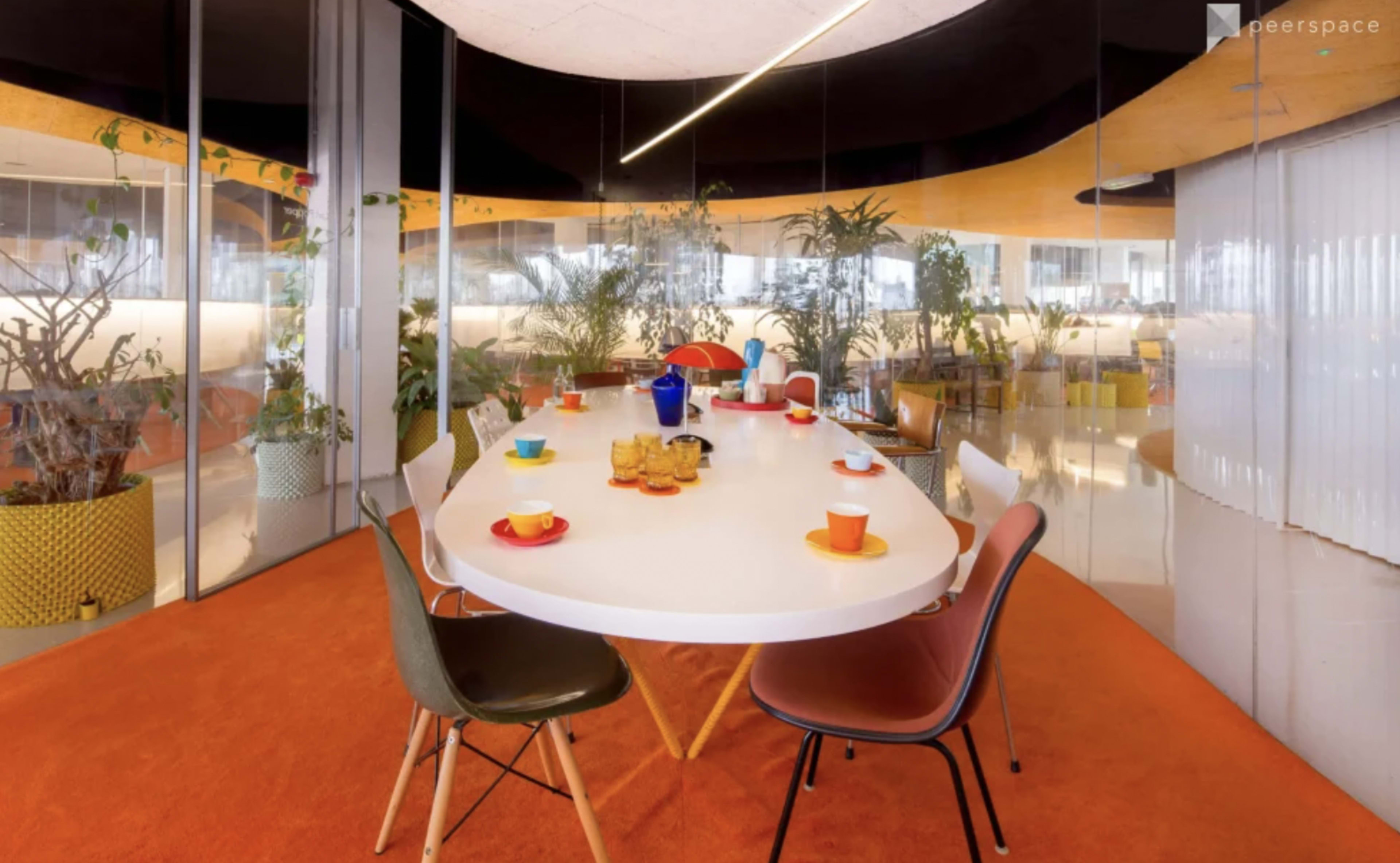 A modern dining area features a large white table surrounded by colorful chairs, with plants and decorative elements in the background.