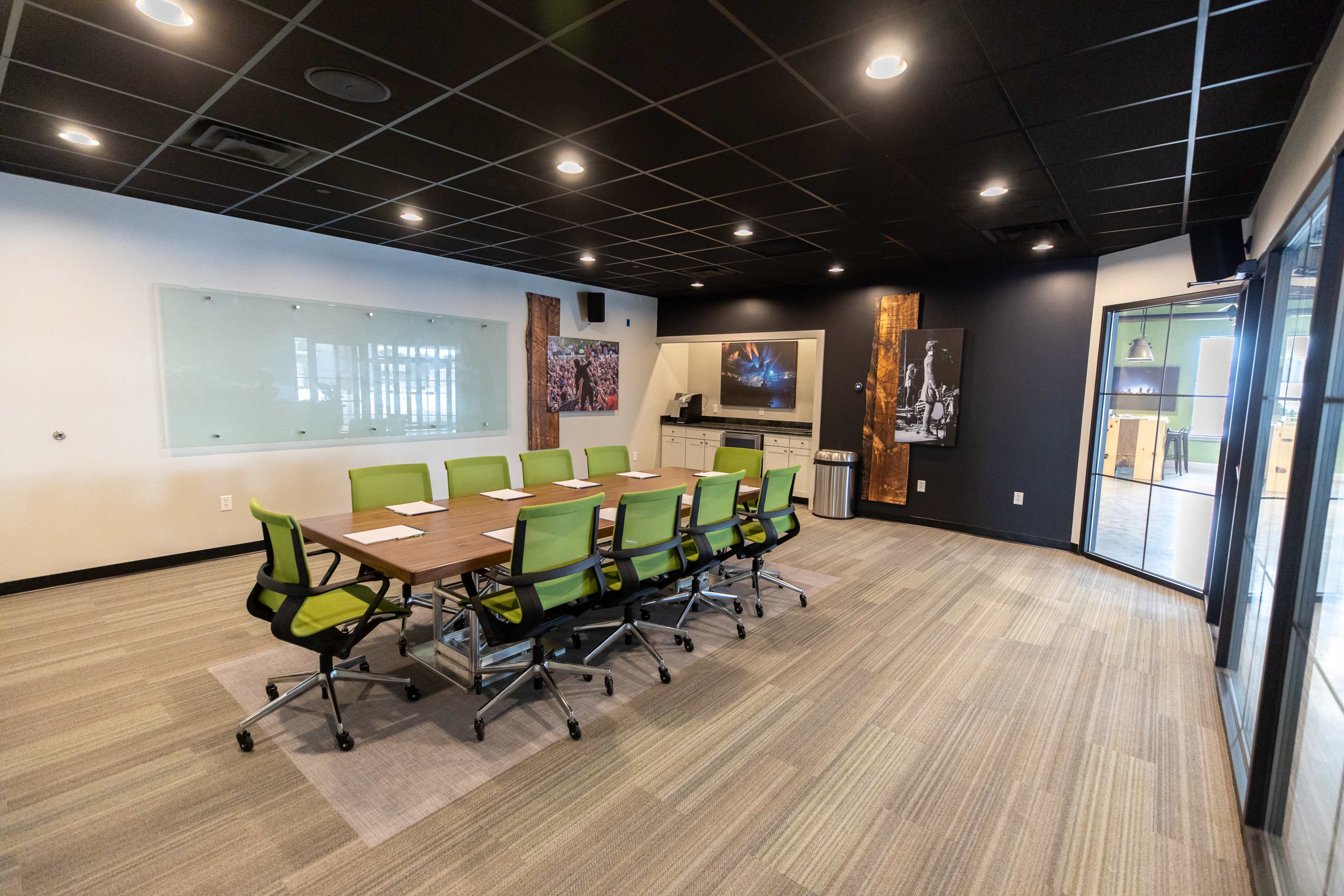 A conference room features a long wooden table surrounded by green office chairs, with a glass whiteboard and artwork displayed on the walls.