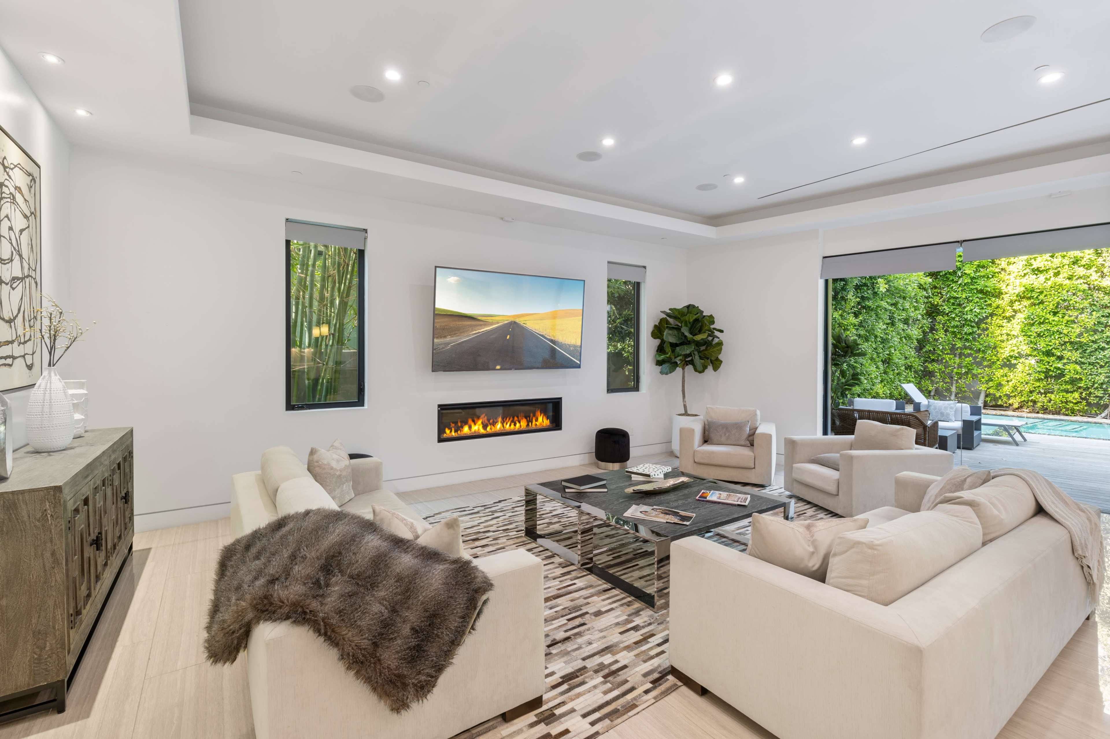 The image shows a modern living room with two light-colored sofas, a coffee table, a large flat-screen TV mounted on the wall, and an electric fireplace beneath it, with windows revealing an outdoor view of greenery and a pool area.