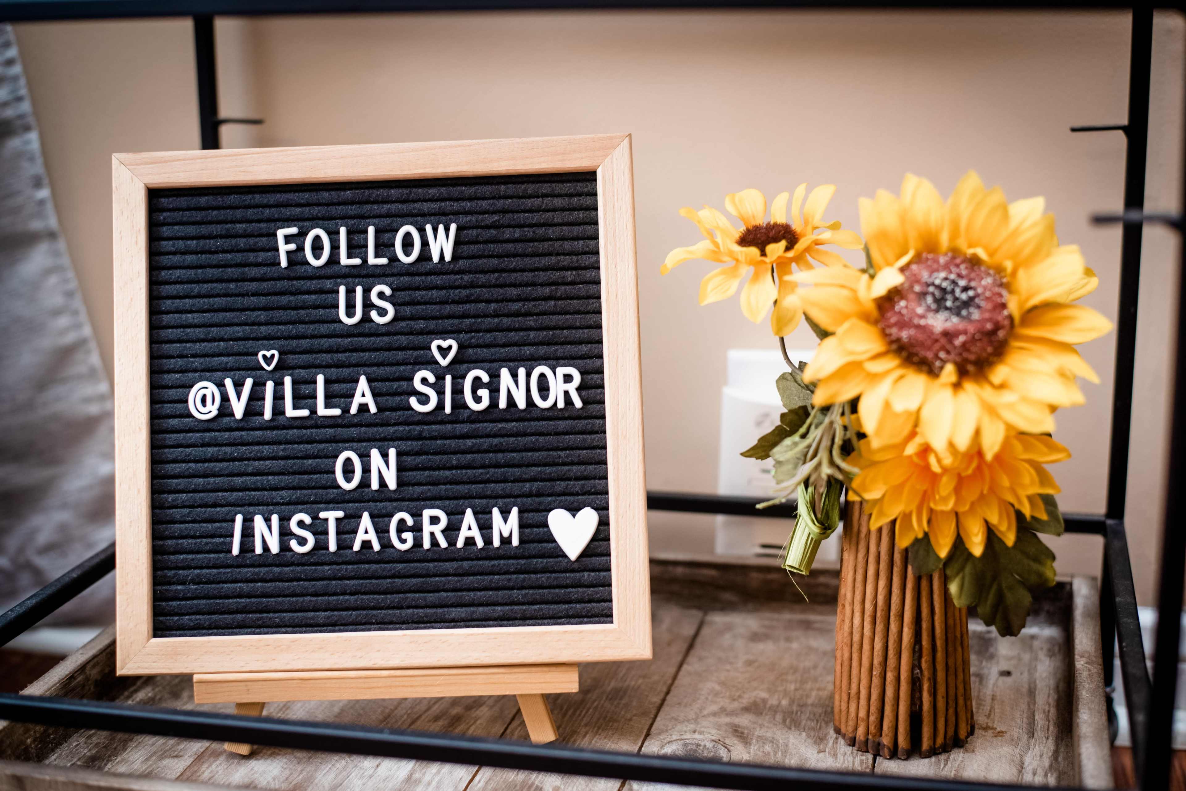 A wooden frame displays a letter board with the text "FOLLOW US @VILLA SIGNOR ON INSTAGRAM" alongside a vase of sunflowers.