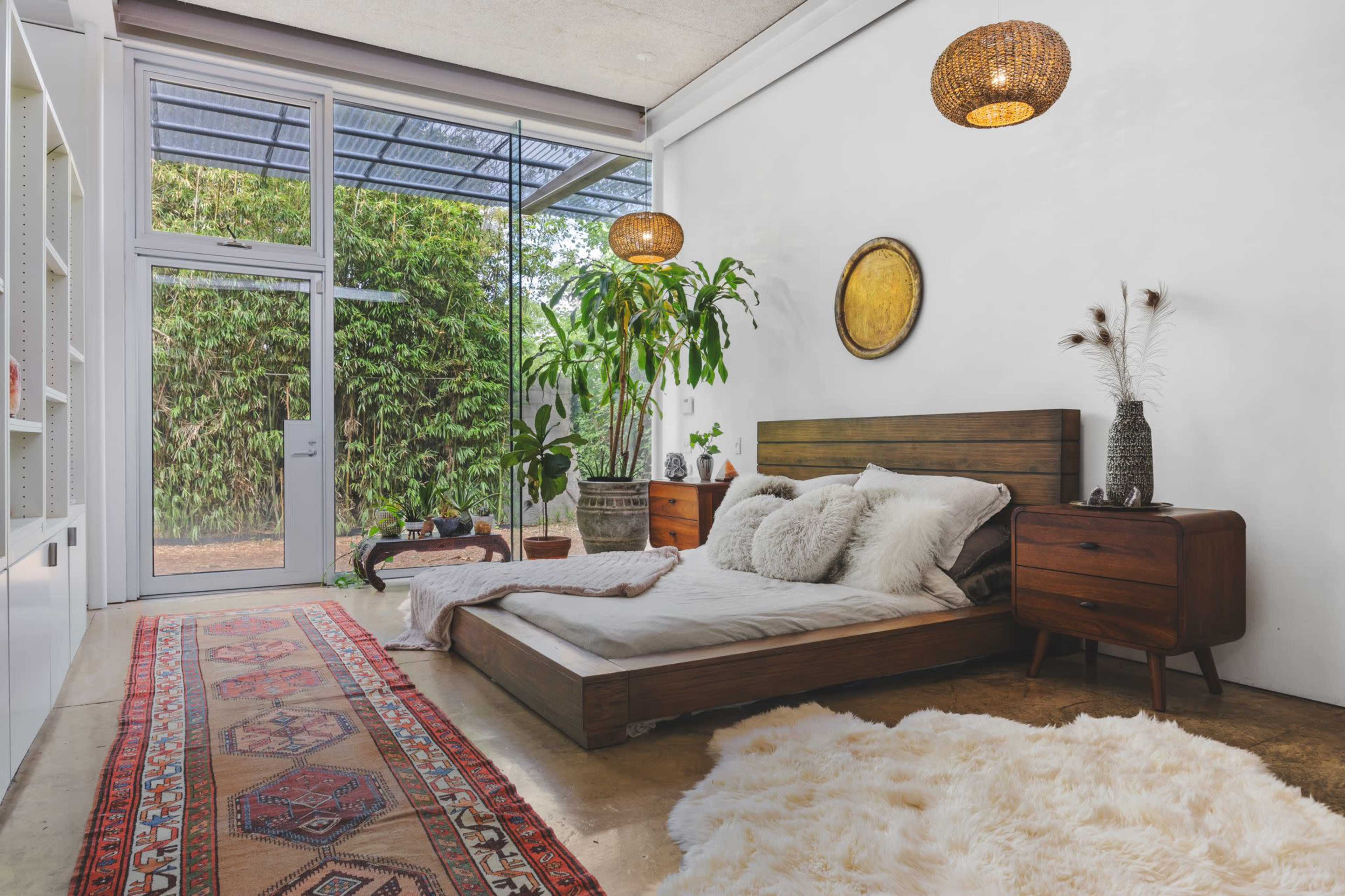 A spacious, minimalist bedroom features a wooden bed with light bedding, a patterned rug, and greenery visible through a large glass door.