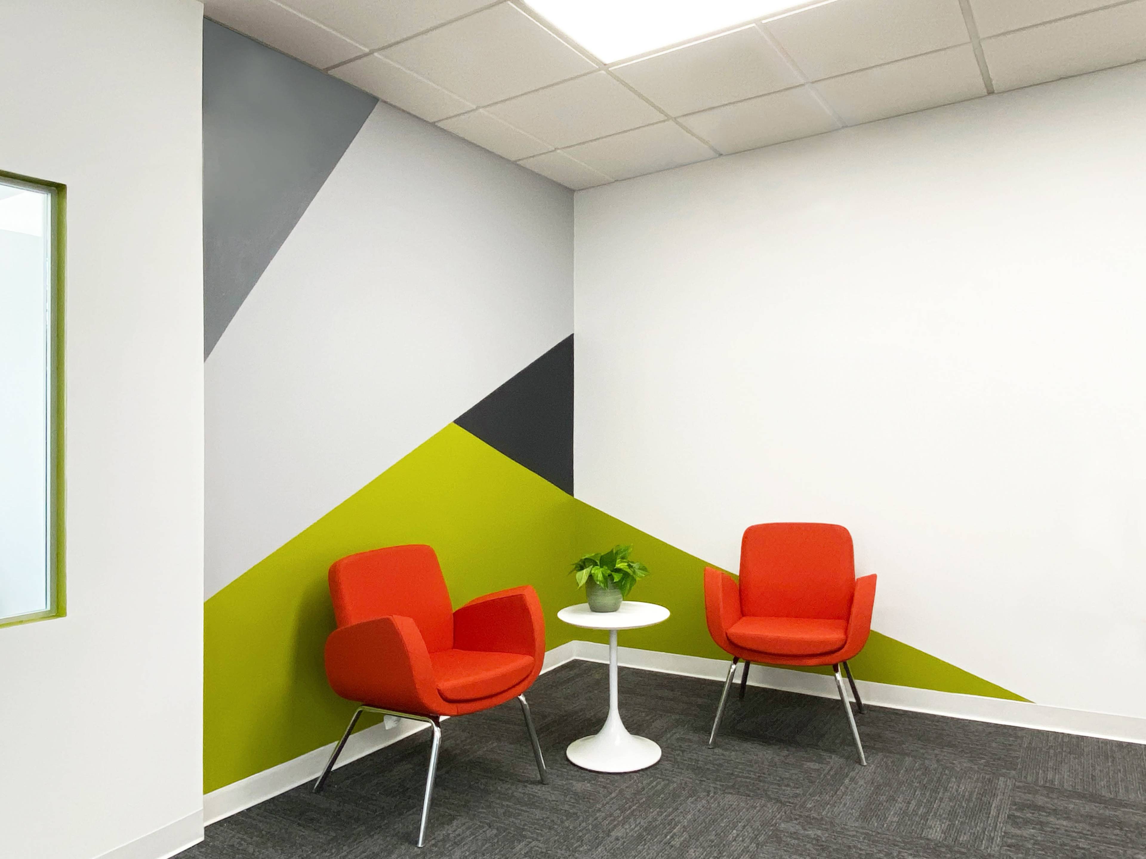 A small seating area with two red chairs and a round white table, complemented by a wall featuring angular geometric painted designs in gray, green, and black.