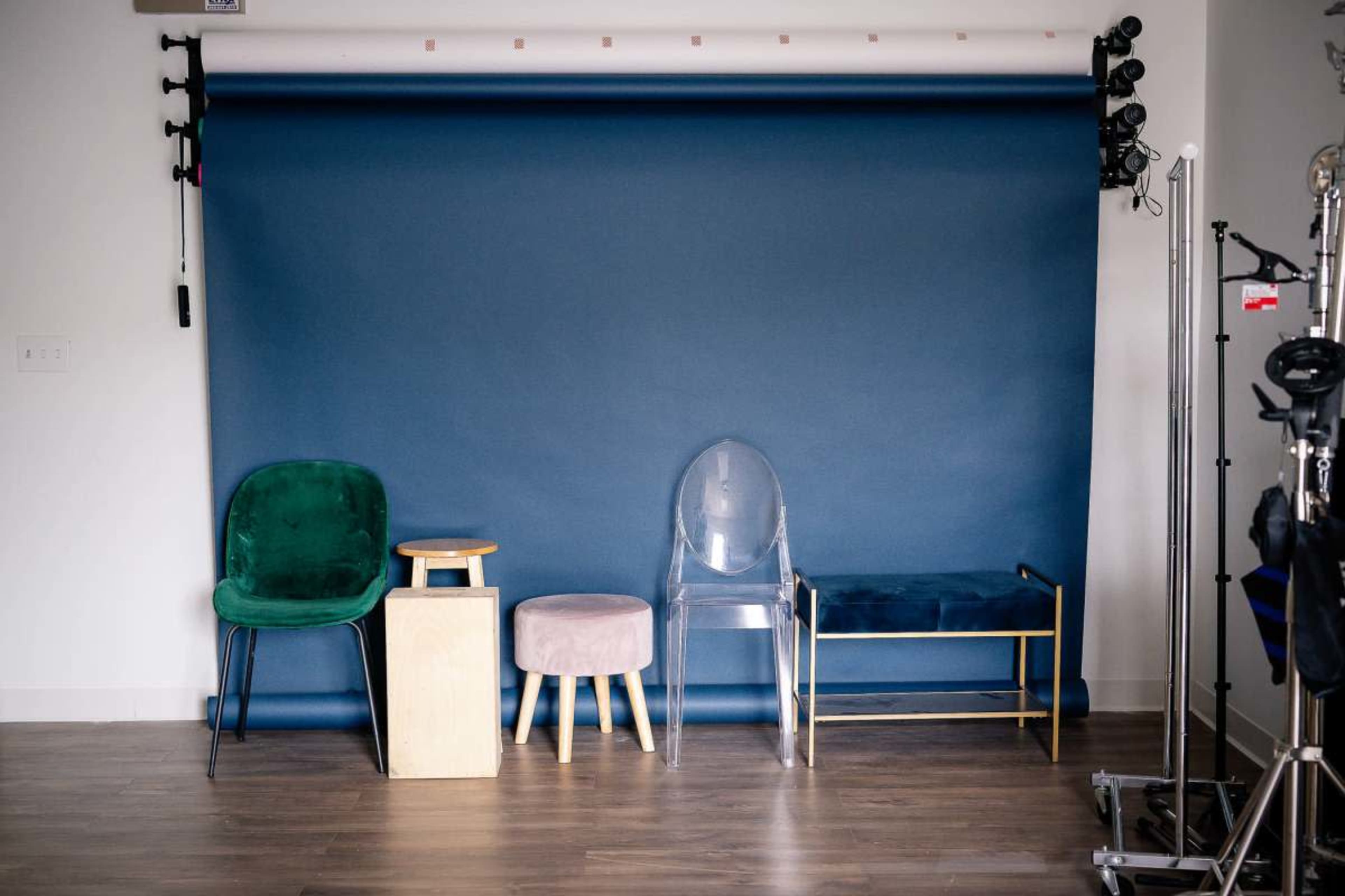 The image shows a row of four chairs, including a green upholstered chair, a wooden stool, a pink velvet ottoman, and a transparent acrylic chair, against a blue backdrop in a studio setting.