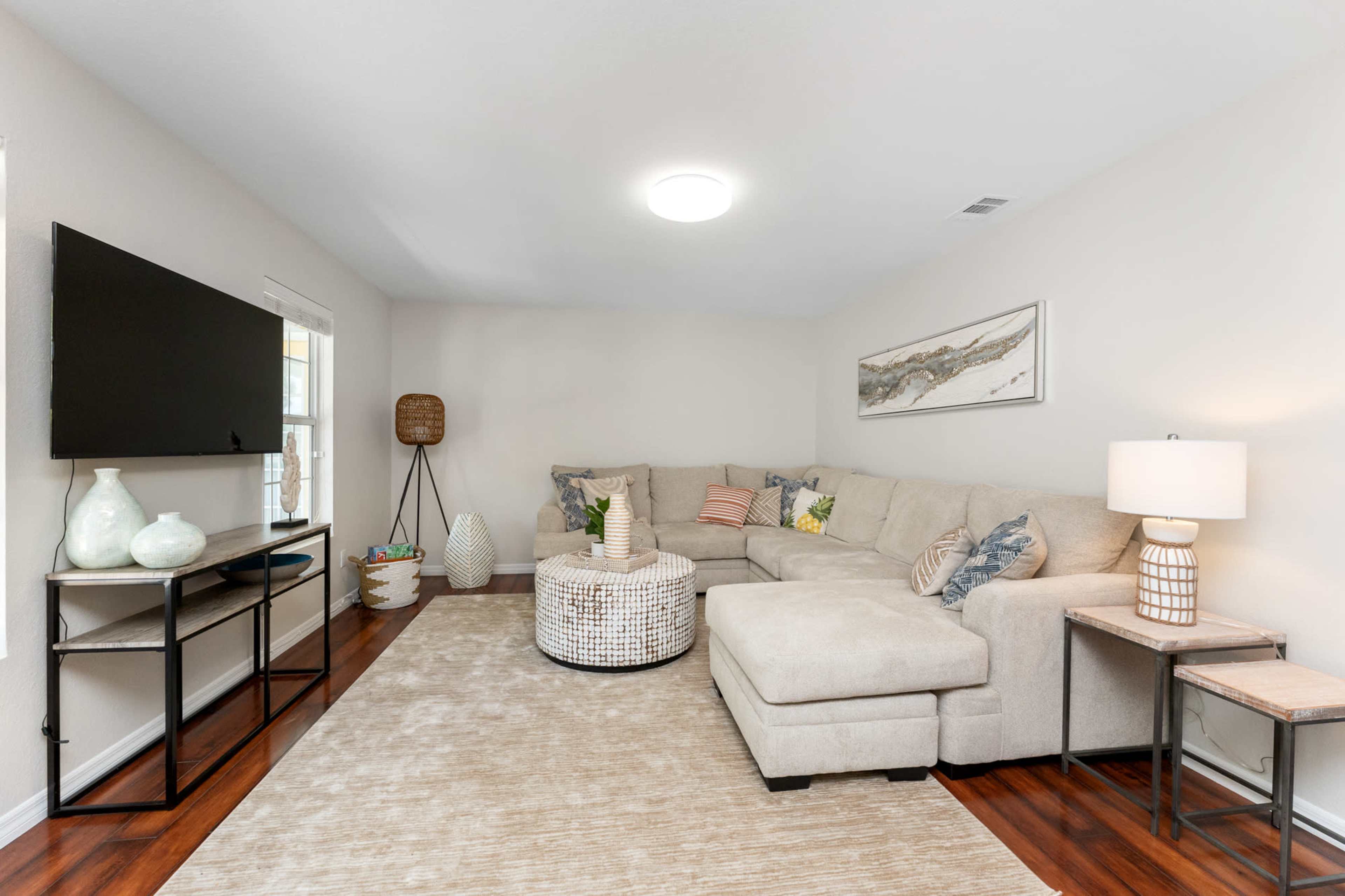 The scene features a light-colored living room with a sectional sofa, a round coffee table, a television mounted on the wall, and a lamp beside a side table.