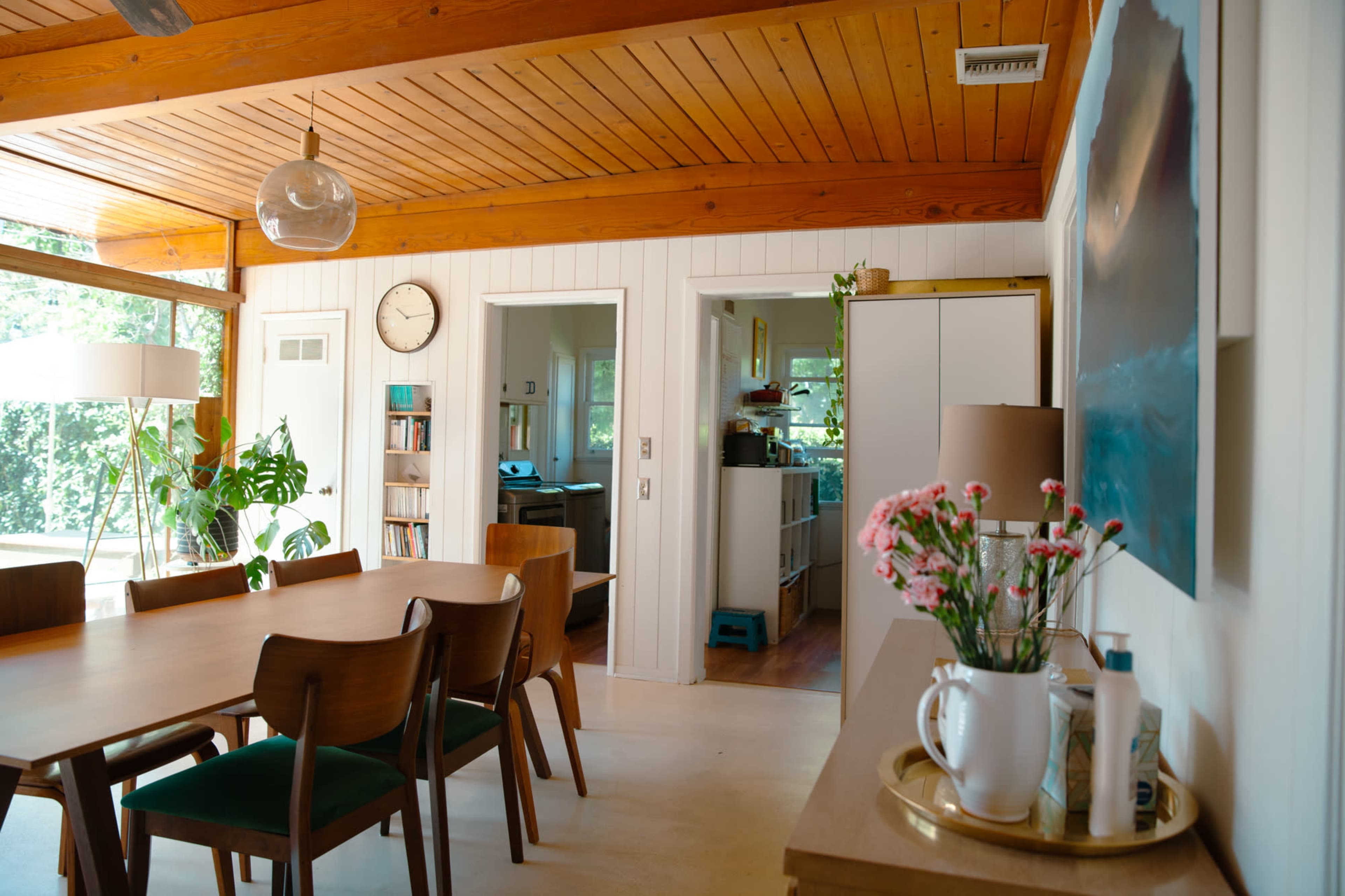 A dining area with a wooden table and chairs, adjacent to a kitchen and a living space featuring a large window and plants.