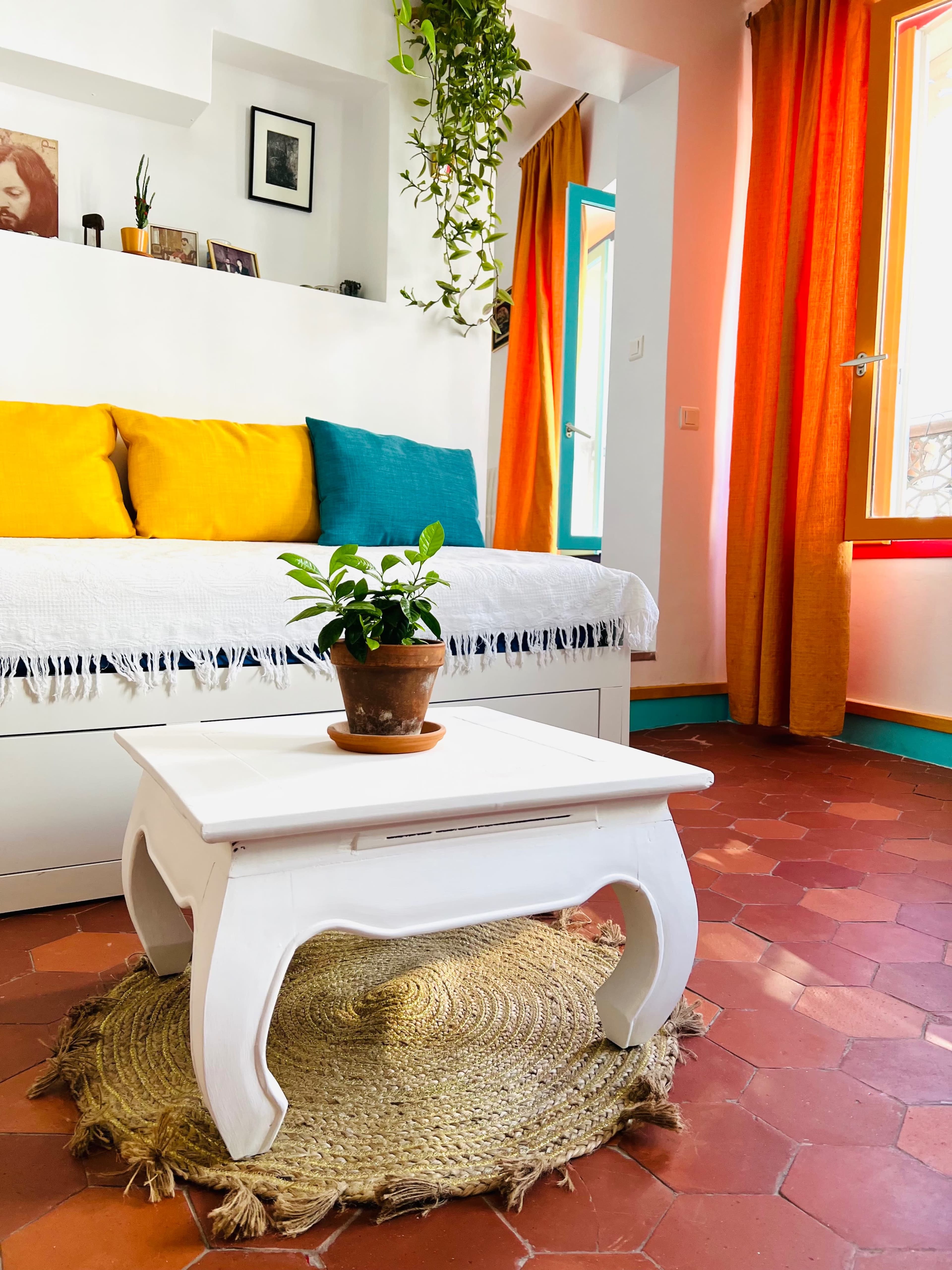 A cozy living room features a white coffee table on a woven mat, with a potted plant, colorful cushions on a daybed, and bright orange curtains.