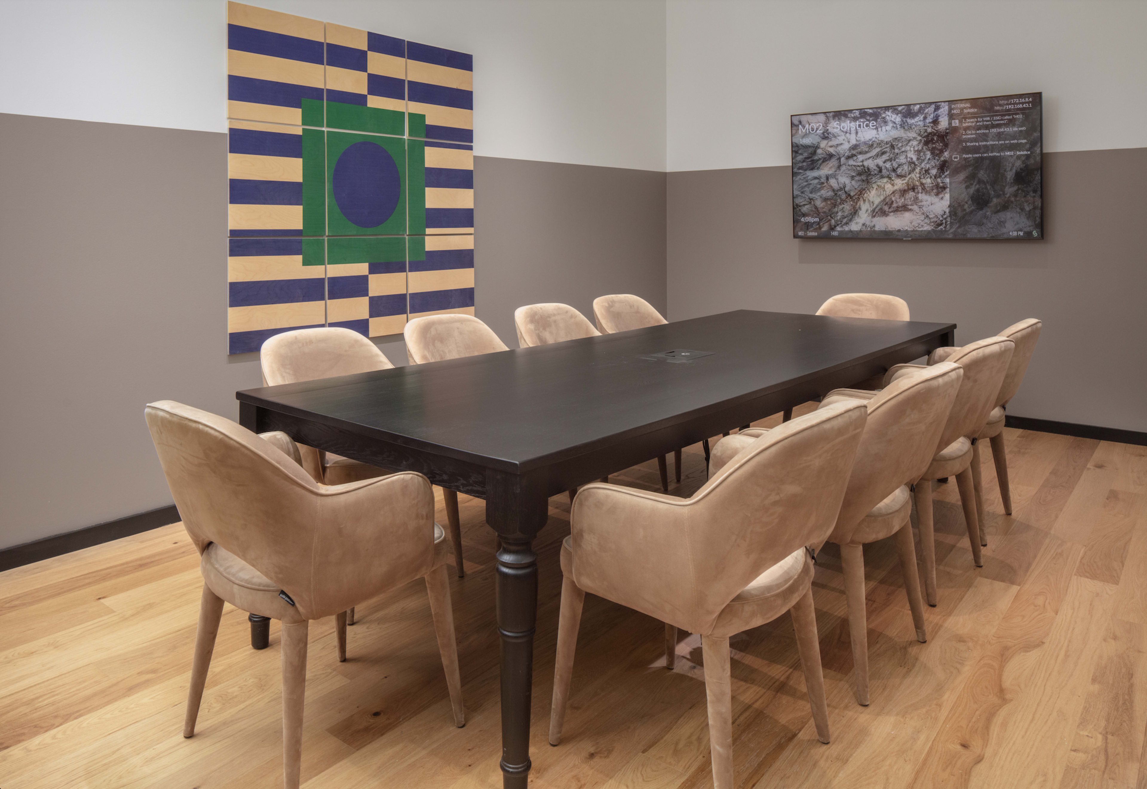 A modern conference room featuring a long black table surrounded by upholstered chairs, with an abstract wall art piece and a screen displaying information on the adjacent wall.