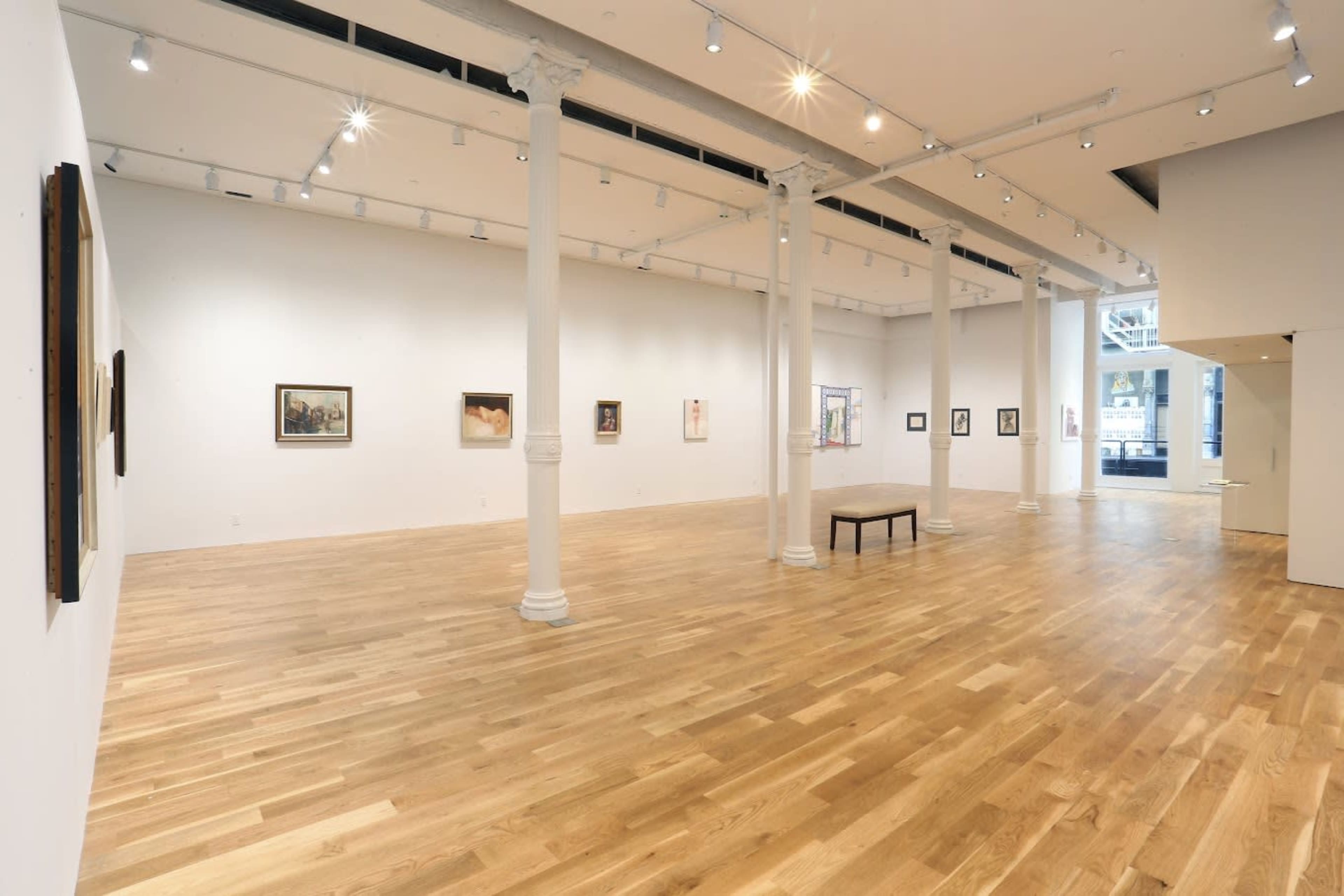 The image shows a modern art gallery featuring wooden flooring, white walls, and several paintings hung along the walls, with a central table placed in the open space.