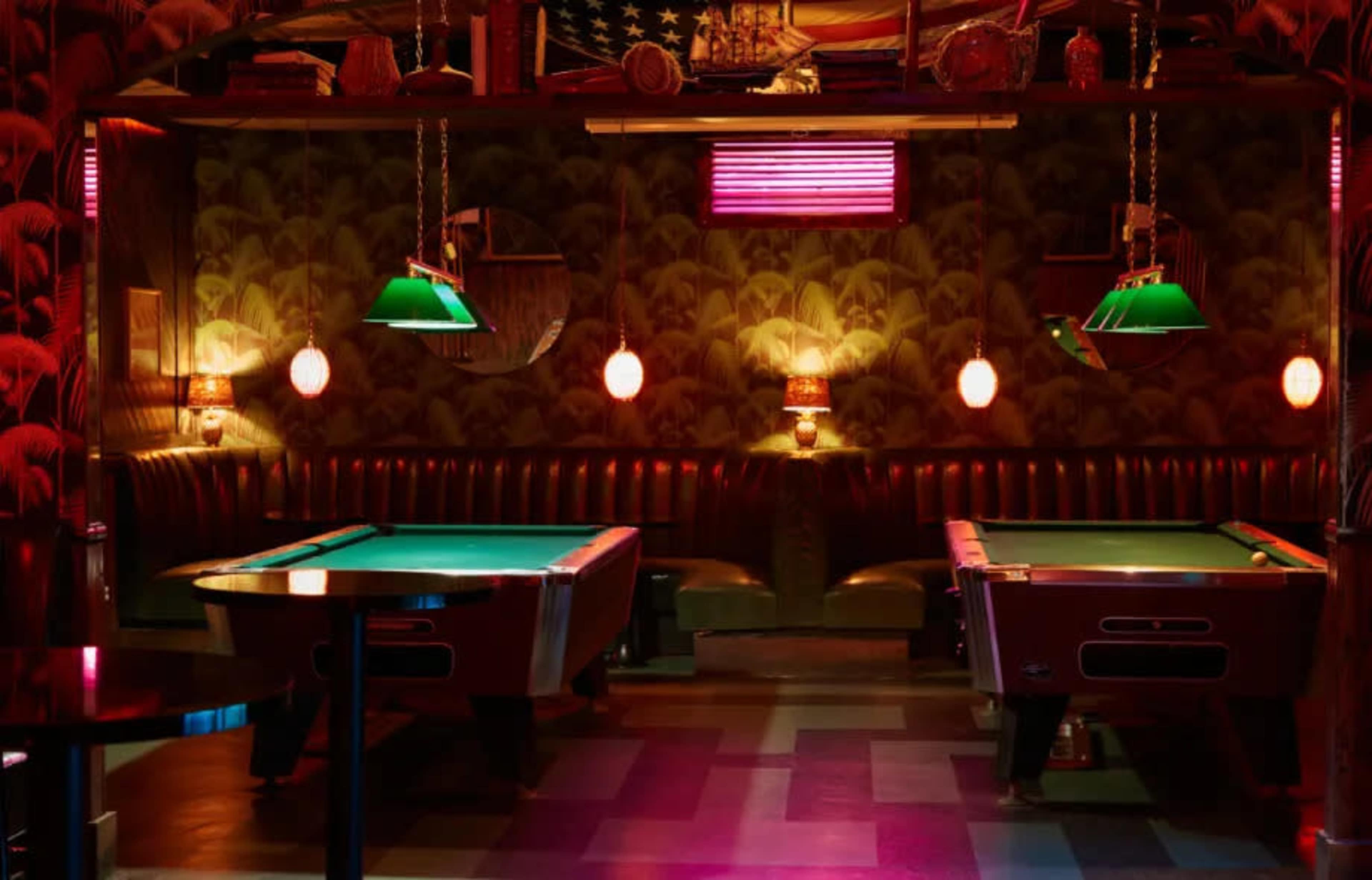 The image shows a dimly lit pool hall with two green-felted tables, vintage decor, and illuminated with colorful lights.