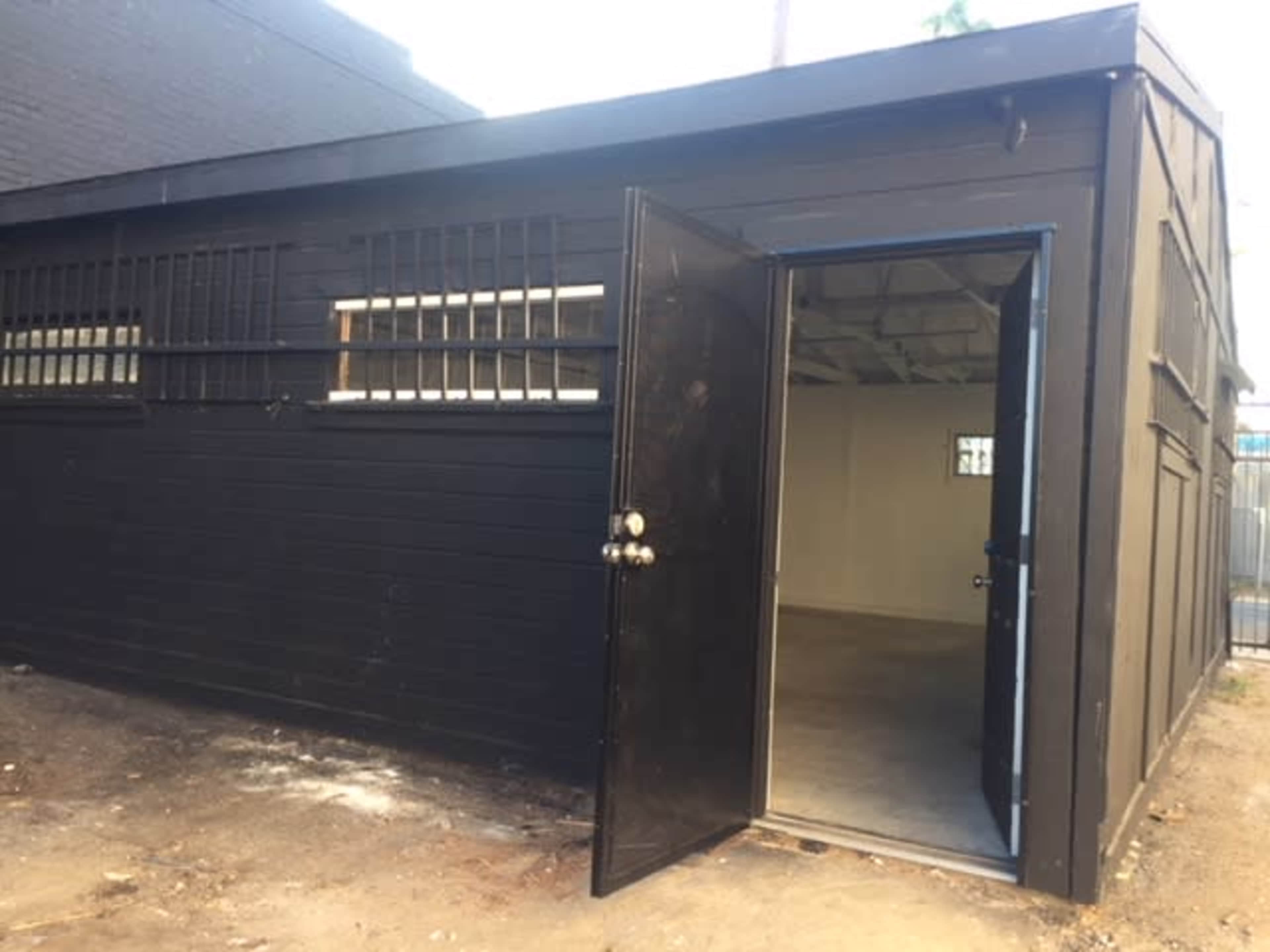 The image shows a black wooden building with an open door and barred windows, revealing an empty interior space.