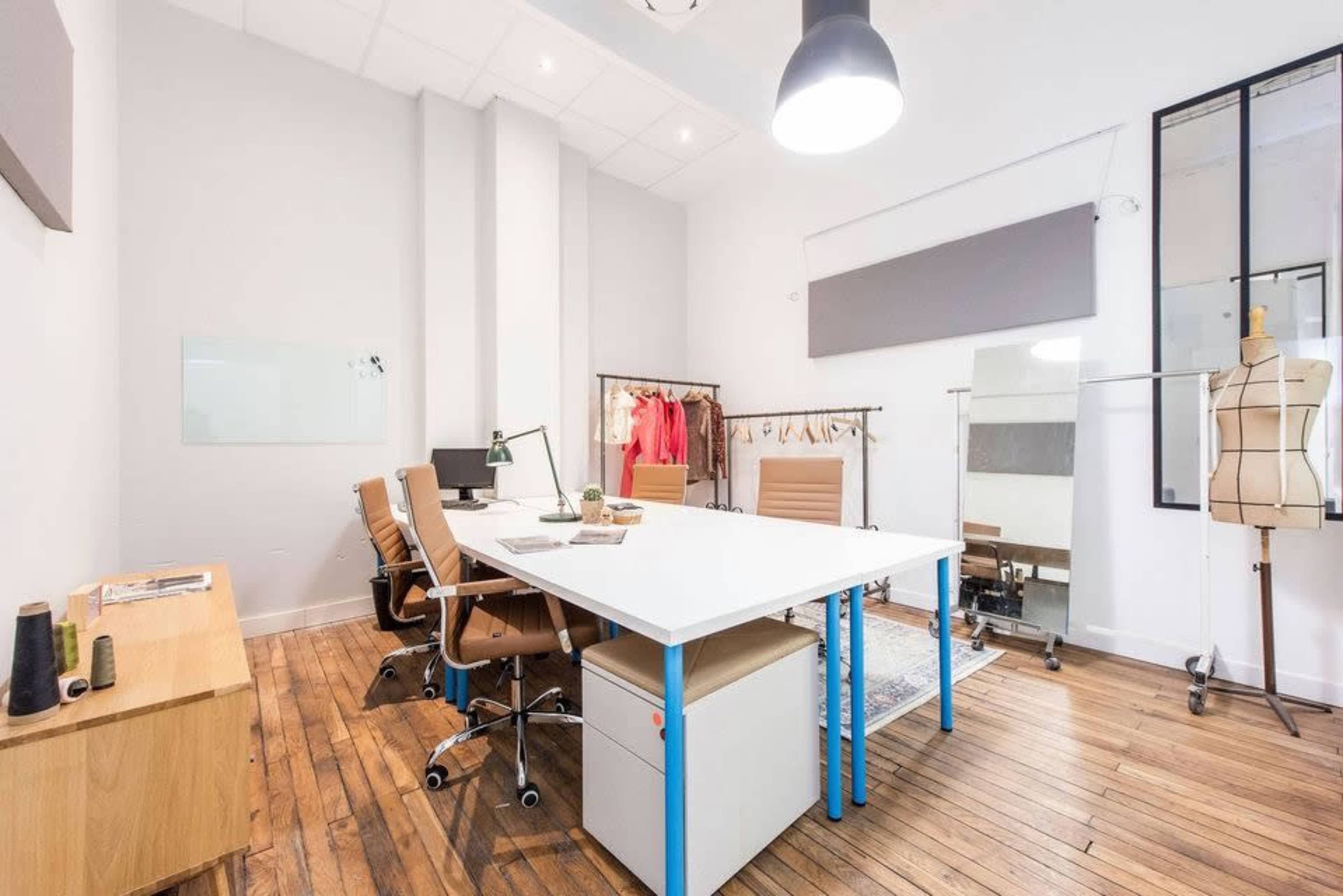 The image shows a modern office space with a large white table, several brown office chairs, and a clothing rack in the corner, all set against a light-colored wall and wooden floor.