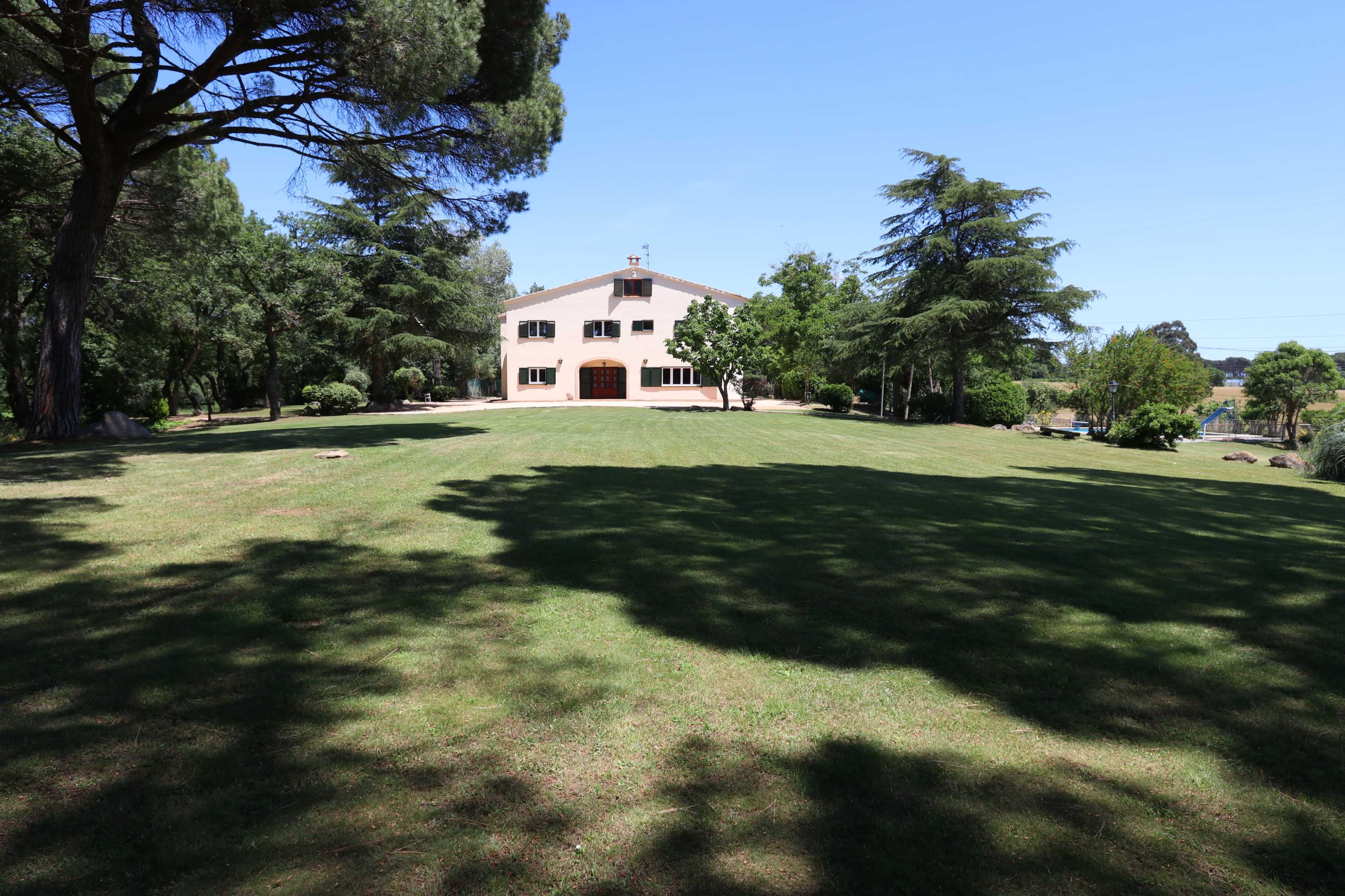 A large house with multiple windows stands amidst a grassy area surrounded by trees under a clear blue sky.