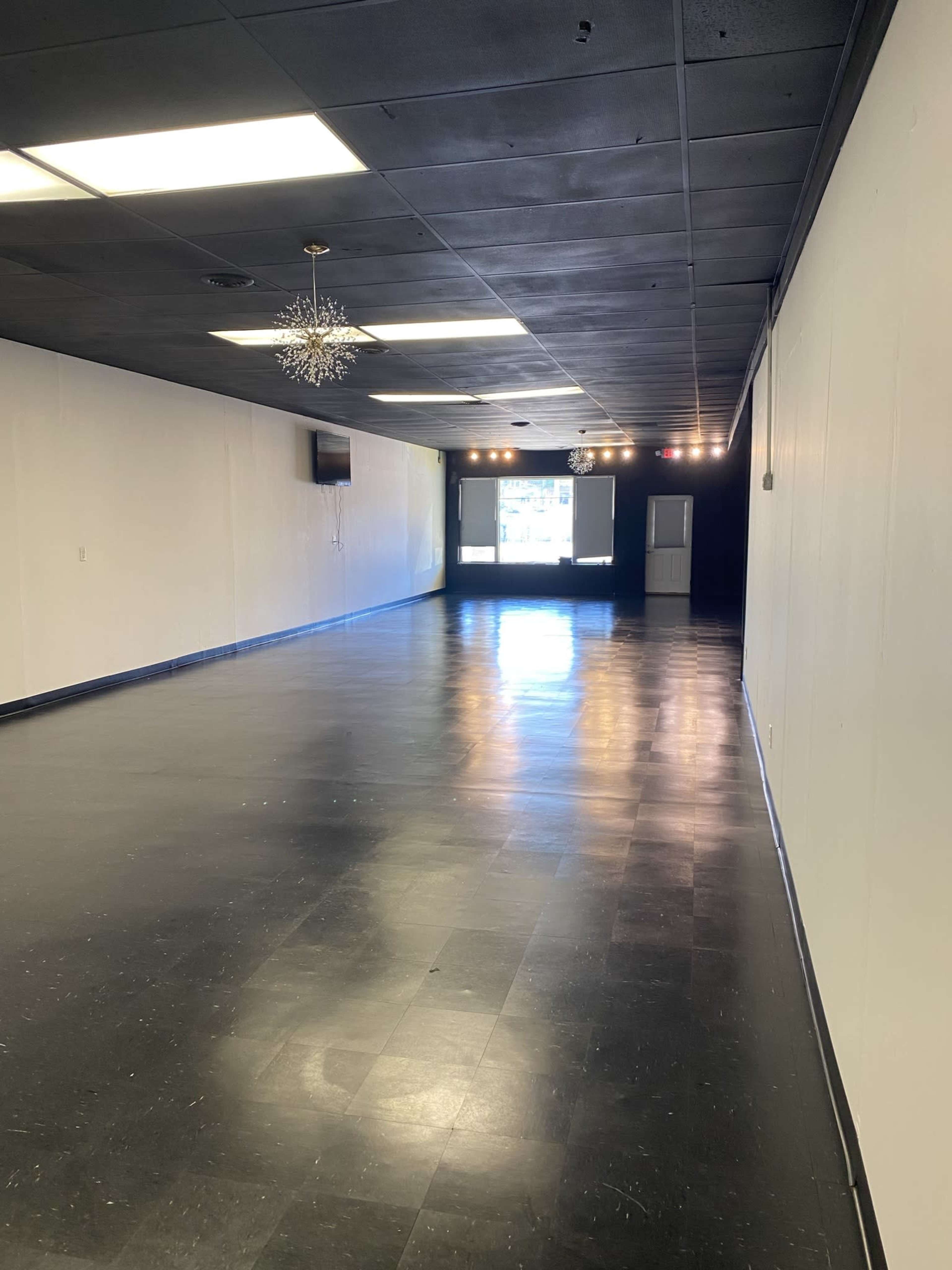 The image shows a spacious, empty room with black tile flooring, white walls, and an illuminated window at the far end.