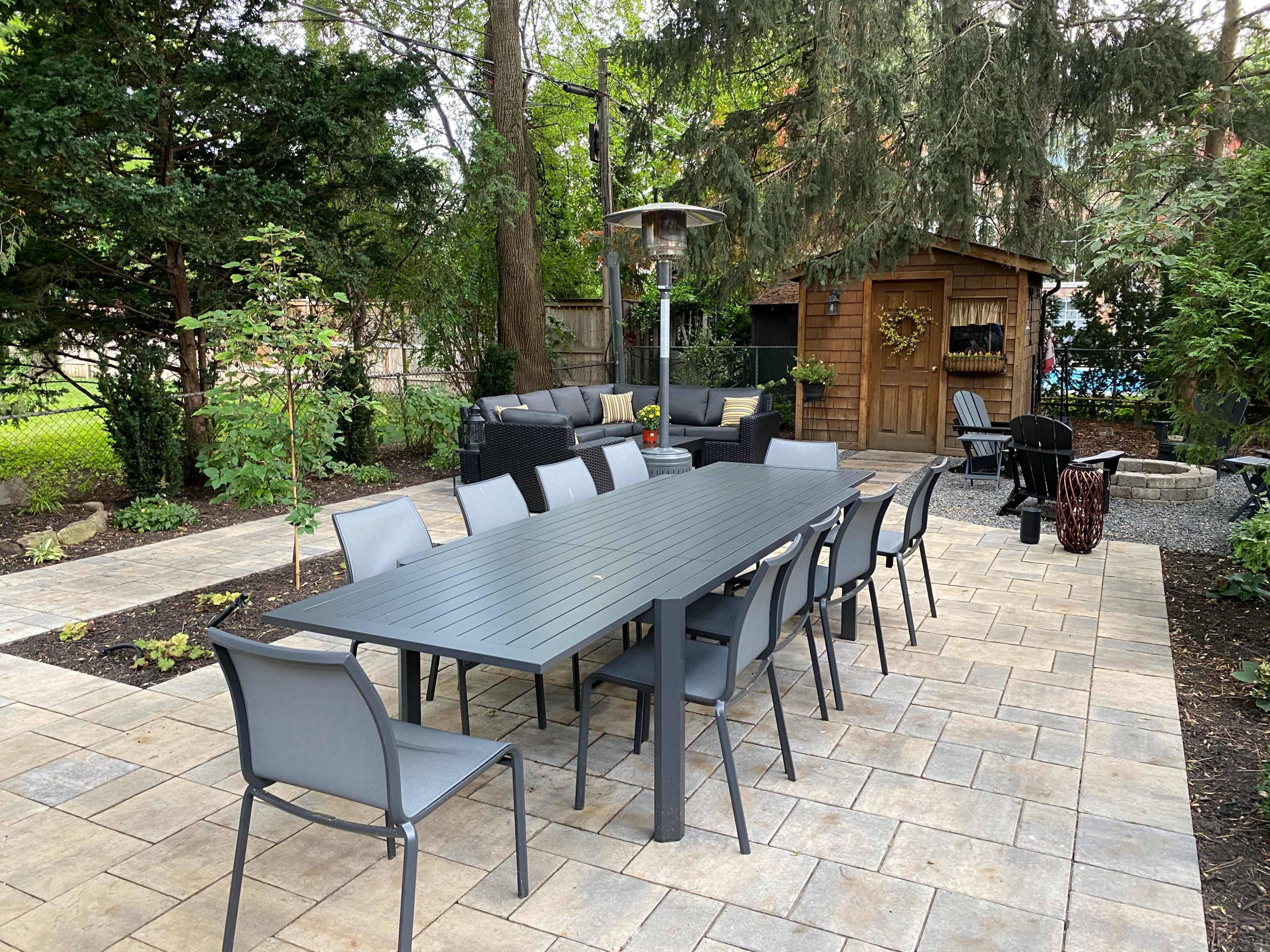The image shows an outdoor patio area with a long dining table surrounded by chairs, a seating area, a patio heater, and a wooden shed in the background.