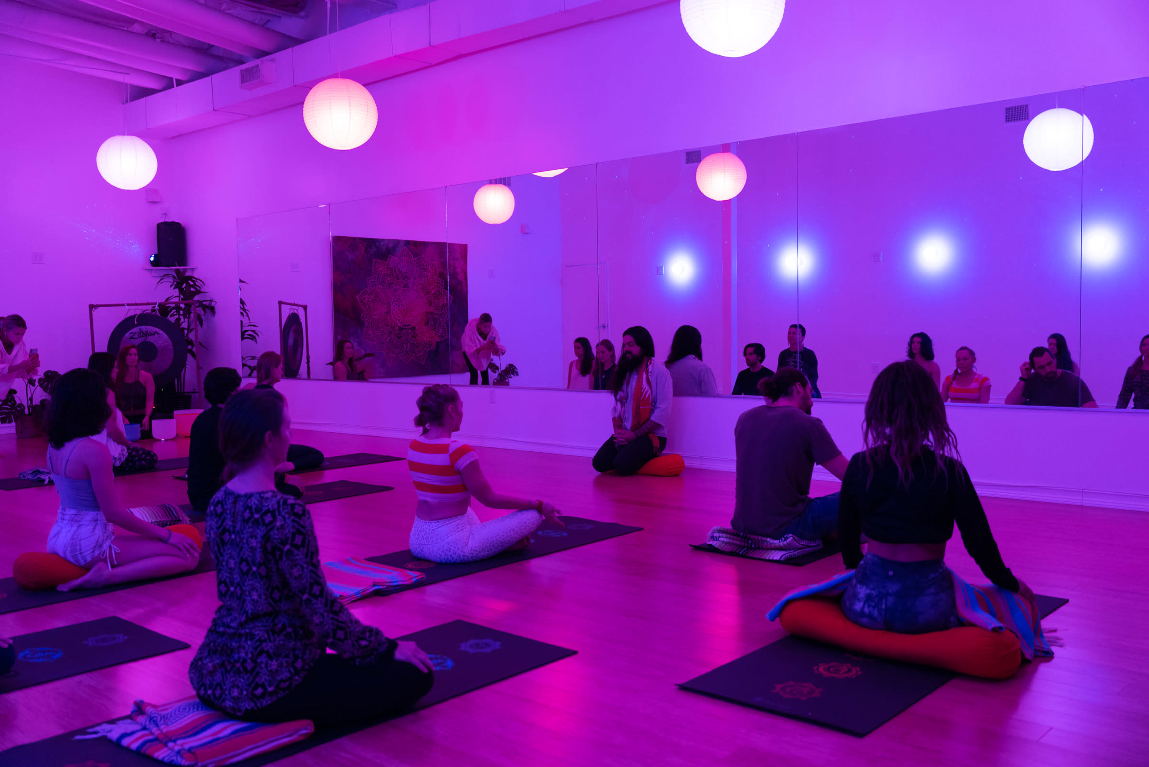 A group of people sits on mats in a brightly lit studio with purple ambient lighting and mirrors, engaged in a yoga or meditation session.