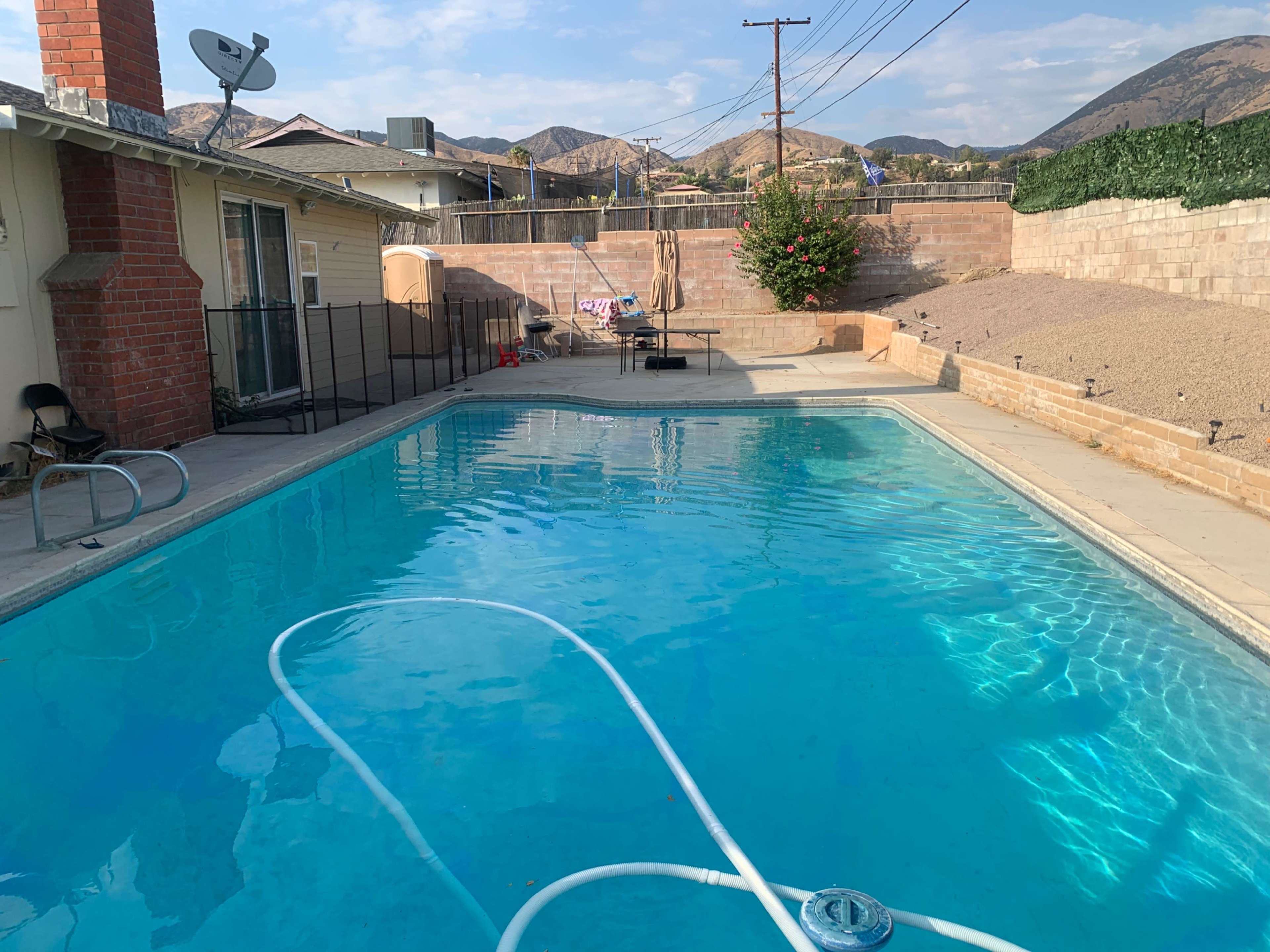 The scene shows a clear swimming pool surrounded by a fenced area, with mountains in the background and patio furniture nearby.