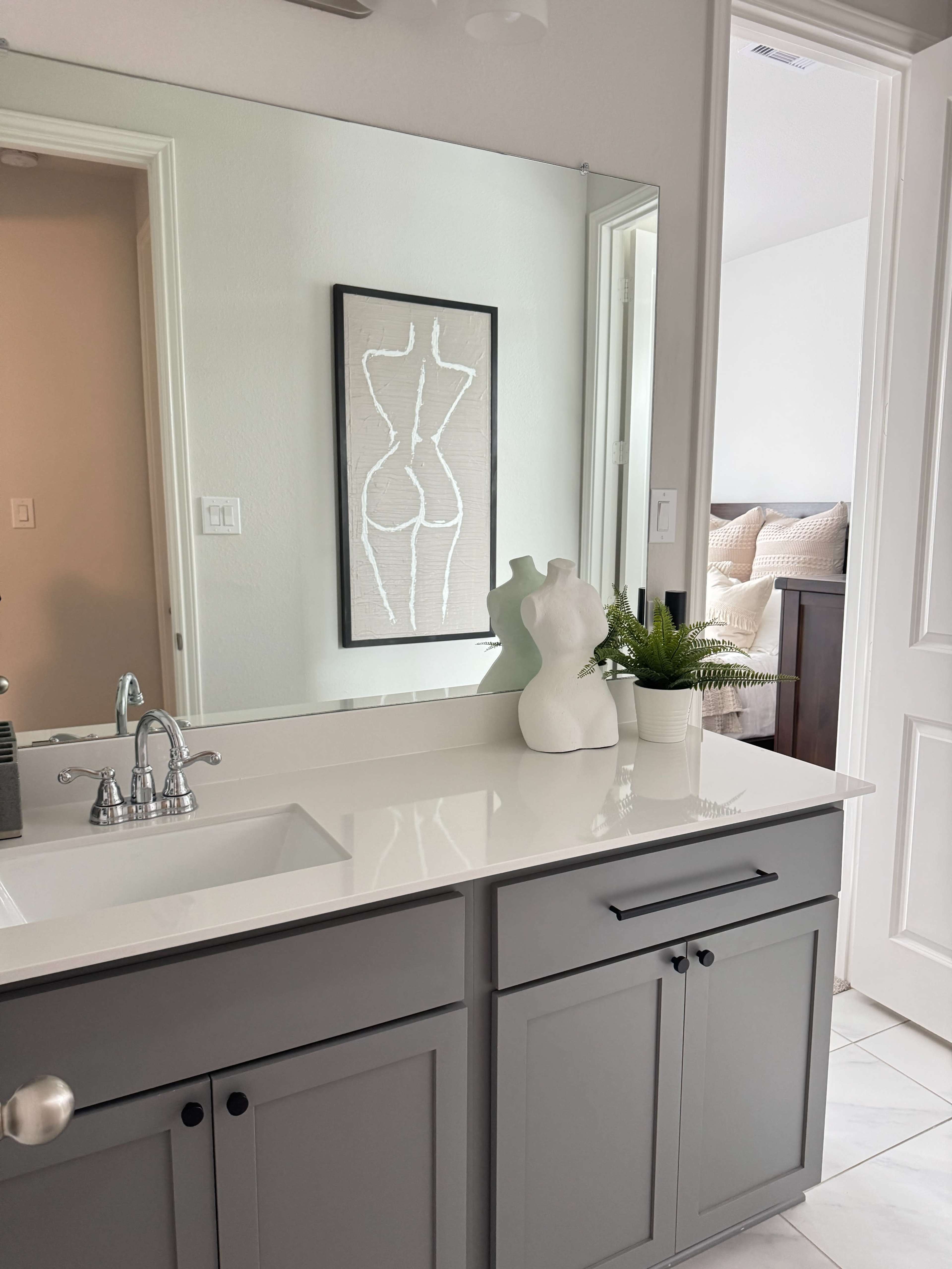 The image shows a modern bathroom vanity with a sink, a framed artistic piece on the wall, a small plant, and a statue against a mirror reflecting an adjacent room.