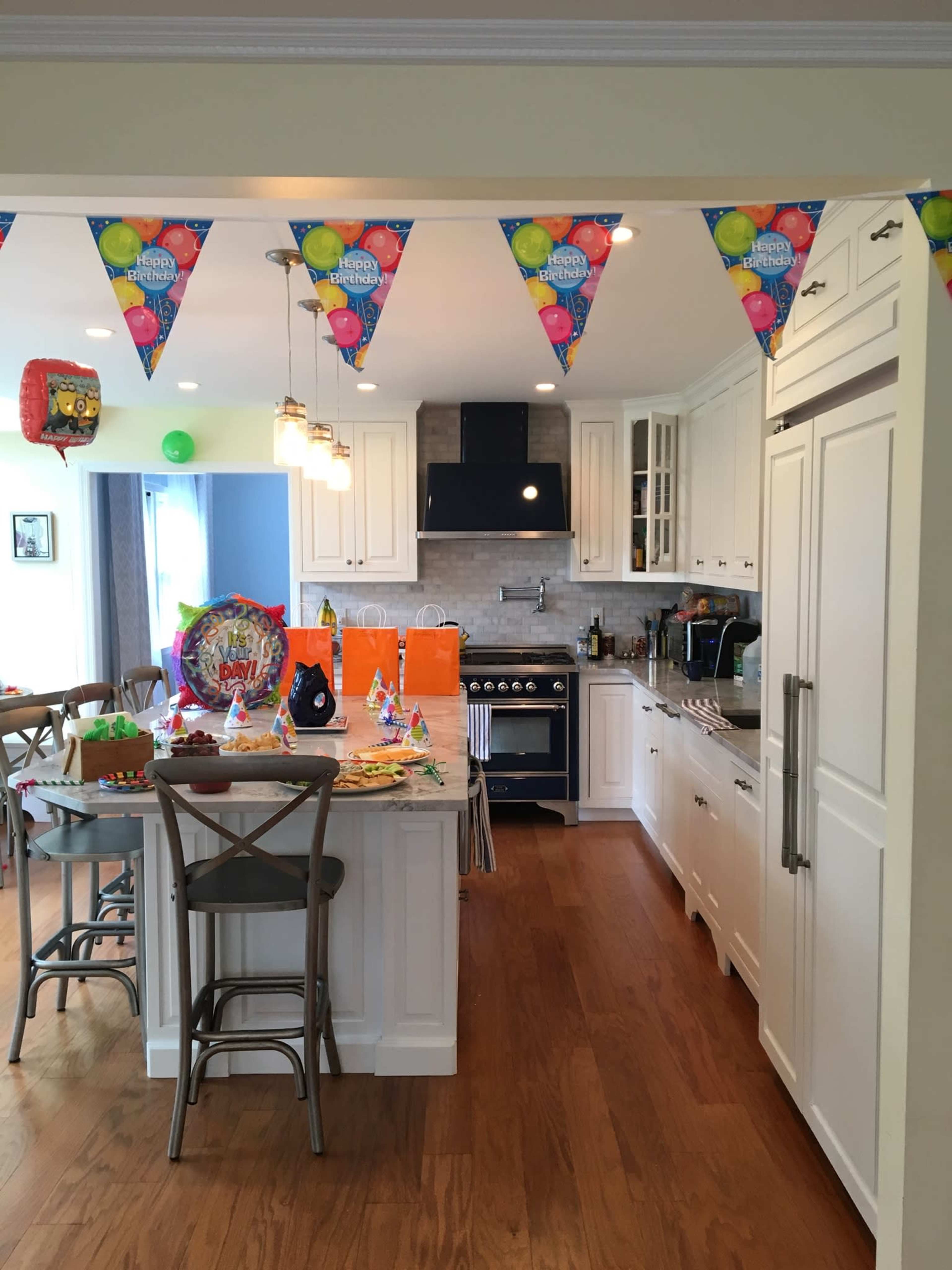 The kitchen features white cabinetry, a large island with stools, and colorful party decorations, including banners and festive tableware.